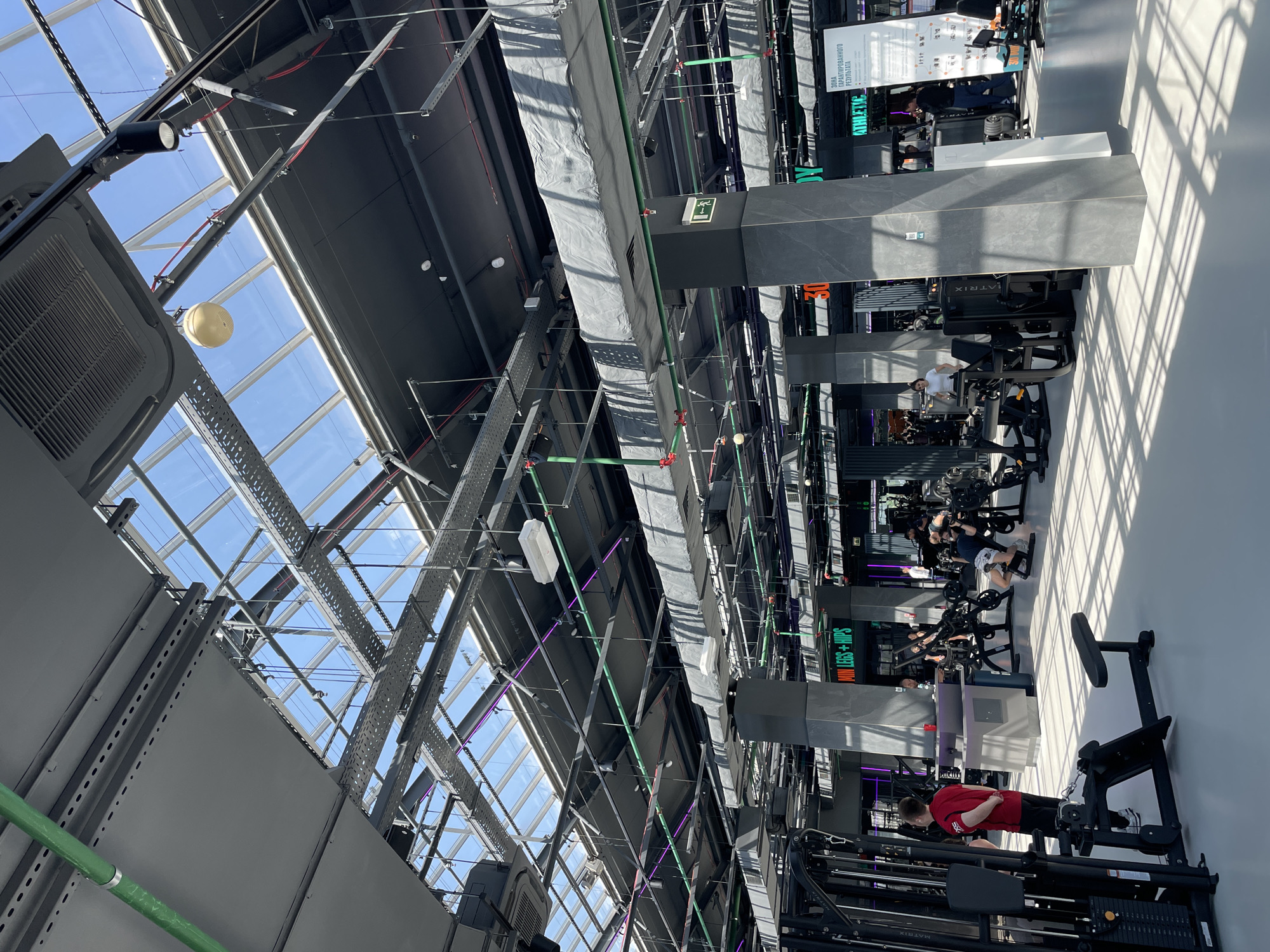 The image captures the interior of a modern gym in Kazan, Russia. The location features a high ceiling with a structural metal framework and skylight panels providing natural light. A complex network of metal conduits and ventilation ducts runs along the ceiling. Exercise equipment, including weight machines, is neatly arranged in rows. Several people are engaged in various workout activities.
