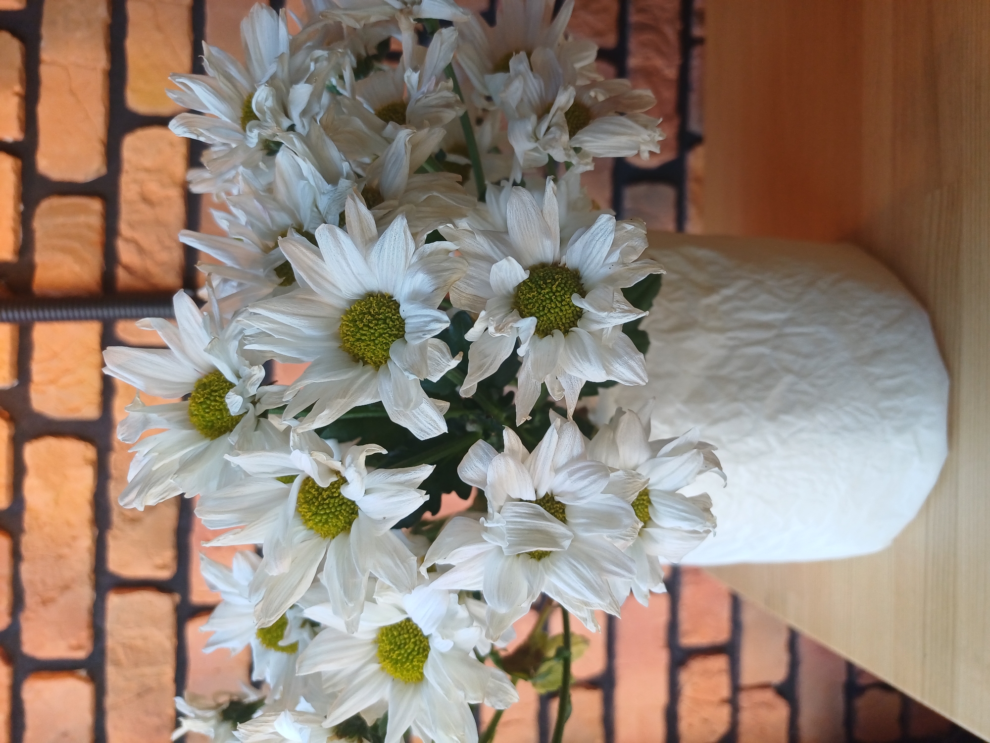 A bouquet of white chrysanthemums with yellow centers is arranged in a white, textured vase. The vase sits on a light brown wooden surface, with a brick wall in the background. The lighting is natural, casting soft shadows. The image is a still life, with no people or animals present. The overall mood is calm and serene. The location in the city of Baryshivka, Ukraine, is not discernible from the image itself.