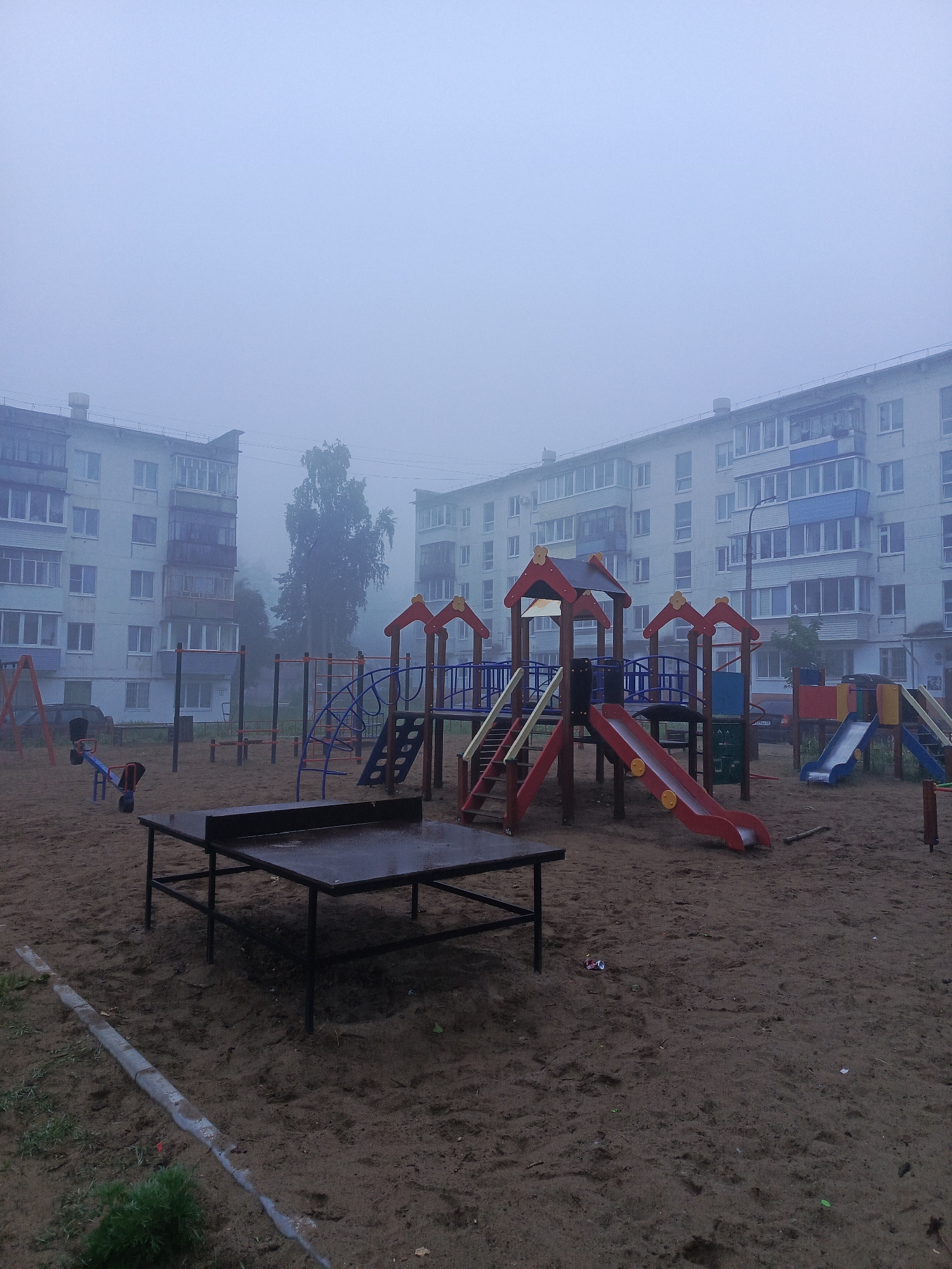 This image captures a deserted residential playground in Chaikovsky, Russia, enveloped in heavy fog. The foreground is primarily sand, with a concrete curb delineating a small grassy patch on the left.
The main subject is a colorful playground structure featuring multiple red slides, blue climbing frames, and small red peaked roofs. To its left, a blue and red seesaw and a partial view of a swing set are visible. A black, wet-looking table tennis table occupies the lower central foreground, suggesting recent rain or heavy condensation.
In the background, multi-story apartment buildings, typical of post-Soviet architecture with numerous balconies, are partially obscured by the dense, light-blue fog. A tall, leafy tree is also visible, shrouded by the atmospheric conditions. The fog creates a low-visibility and ethereal ambiance.
The playground is empty, with no people present, contributing to a quiet and somewhat melancholic atmosphere. The diffused light indicates either early morning or late evening. No discernible text is visible within the image.