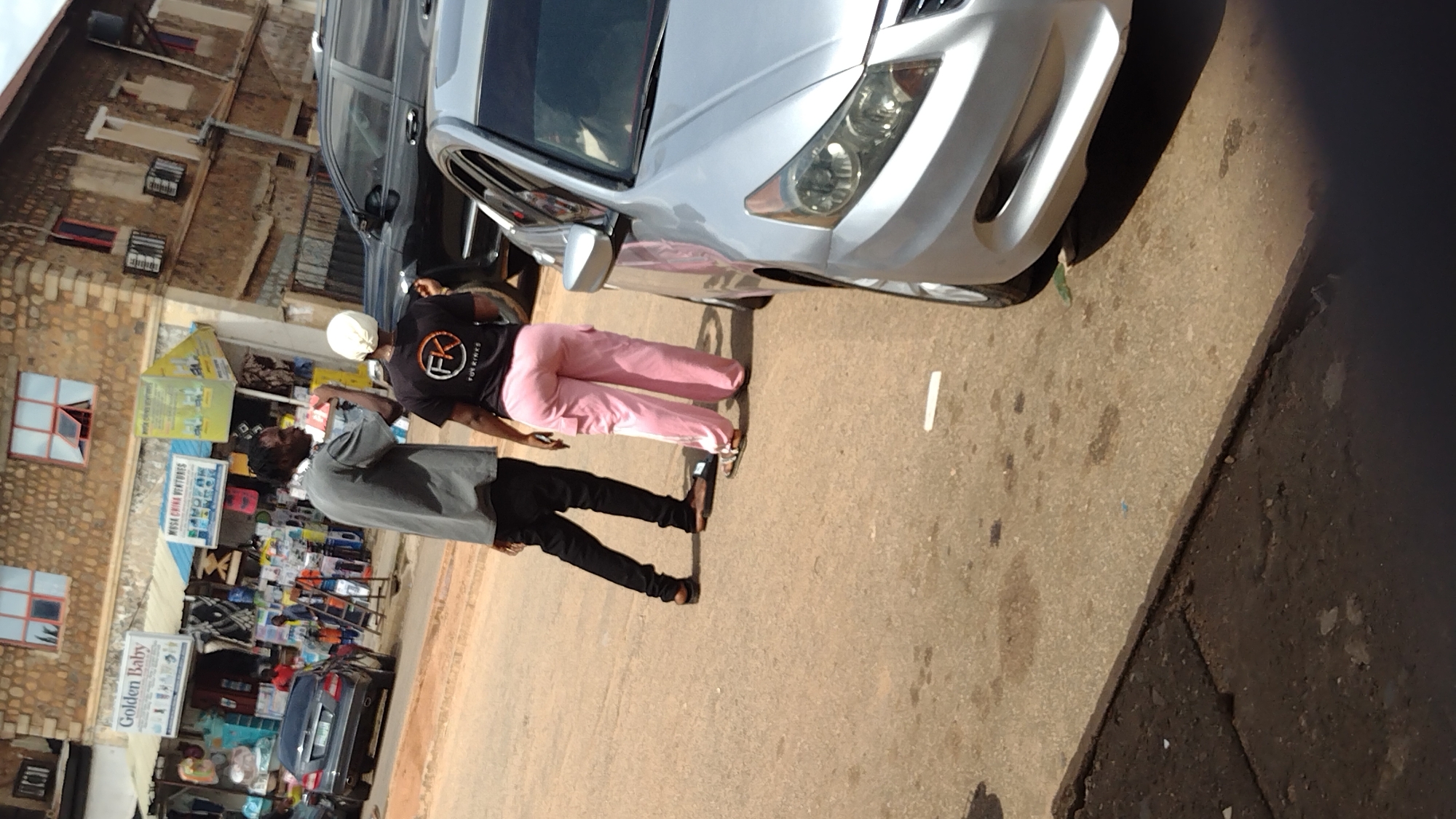 The image depicts a street scene in Jos, Nigeria. Two individuals are the primary subjects. One person, wearing a black t-shirt with a circular logo that reads "FK YOUR VIBES" and light pink pants, stands facing away from the camera. They are also wearing a light-colored head covering. Beside them, a second person, dressed in a grey long-sleeved shirt and black pants, is also facing away from the camera. They are both standing on a paved surface, likely a road or sidewalk, with a silver car parked to their left.
In the background, there are buildings constructed with what appears to be stone or brick. Several windows with red frames are visible on one of the buildings. Adjacent to the buildings, stalls or shops are set up, displaying various goods. Signs with text are present, including one that says "GOLDEN BABY" and another that is partially visible with text that seems to include "WVA CHINA VENTURES."
The lighting suggests it is daytime, possibly late morning or early afternoon, given the strong shadows cast by the car and people. The weather appears to be clear, with ample sunlight. The overall impression is of a bustling market or street-side commercial area. The people's postures don't convey any strong emotions, appearing to be engaged in a casual interaction or simply standing. The presence of parked cars and the shops indicates a functional urban environment.