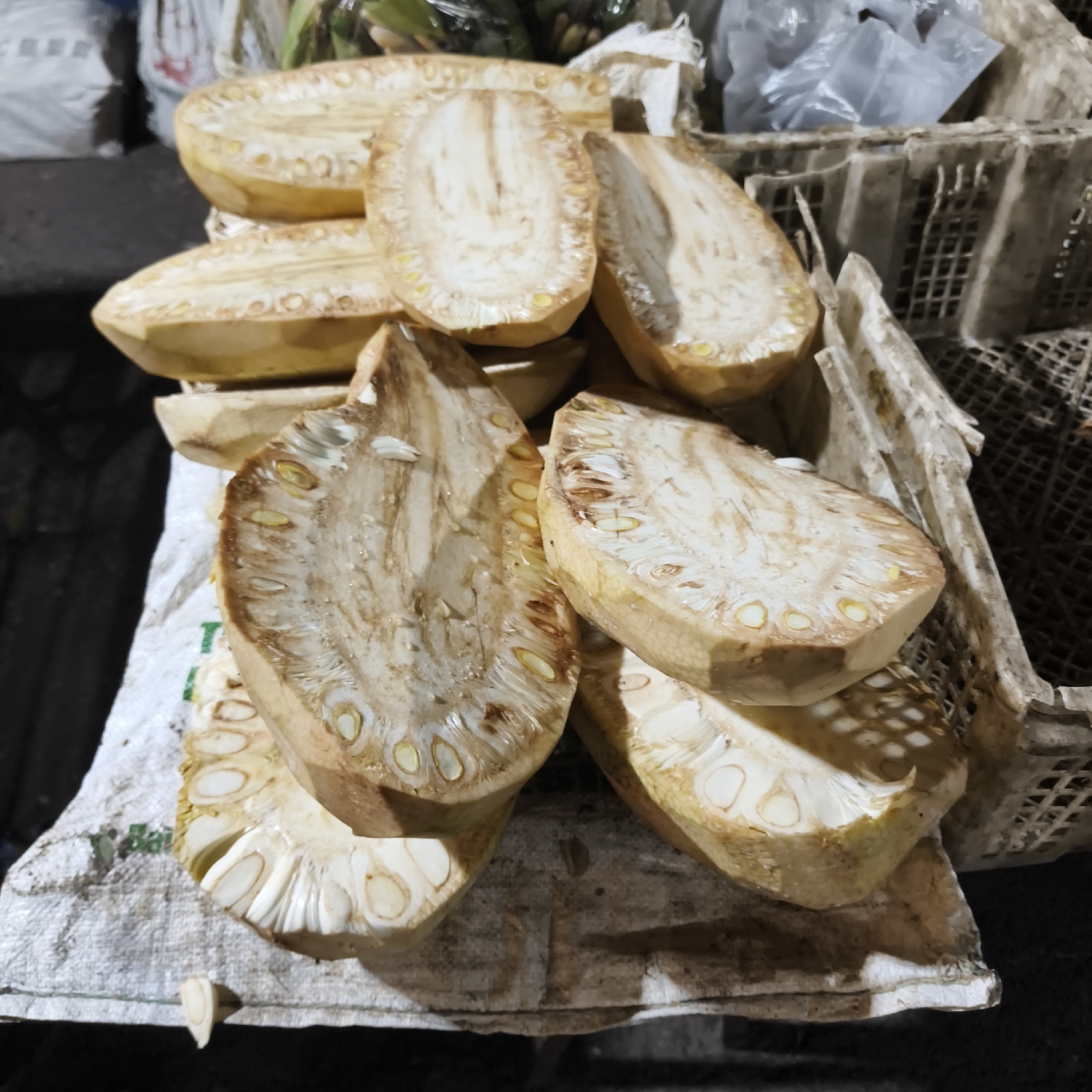 In South Jakarta, Indonesia, sliced jackfruit sections are displayed on a white woven sack. The fruit is a light brown on the exterior, and the interior flesh is a pale yellow with visible seeds. Some pieces are stacked on top of each other, while others are arranged alongside in a woven plastic crate. The background shows more produce and plastic bags, suggesting a market setting.