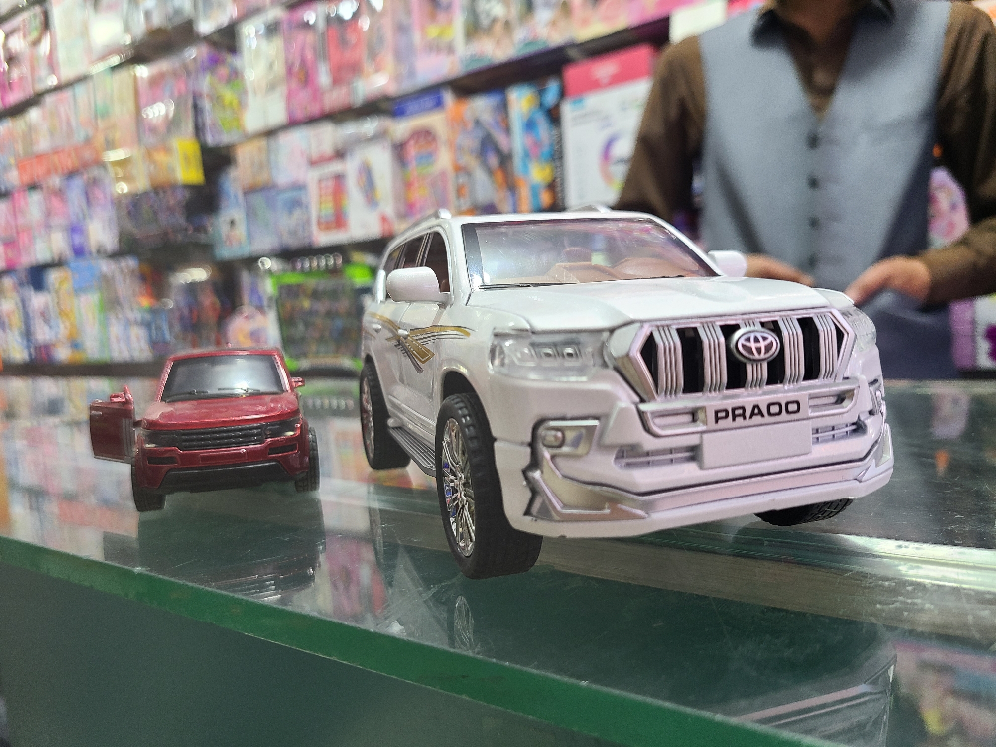 The image depicts a retail environment, likely a shop, situated in Quetta City Tehsil, Pakistan. In the foreground, on a reflective green surface that is possibly a glass display case, are two miniature toy cars. A white toy Toyota Prado with the word "PRADO" prominently displayed on the front grill is positioned closer to the viewer, to the right. A smaller, red toy car with an open door is to the left of the Prado. The toys are displayed on the reflective surface of a glass display case.
In the background, slightly out of focus, shelves packed with various toys and merchandise can be seen. A person wearing a grey vest over a brown long-sleeved shirt stands behind the display case, partially visible and positioned between the toys and the shelves. The person is likely an employee or salesperson at the shop. The shelves are lined with various items, suggesting a toy or general goods store. The presence of the toys and the individual in the background suggests a commercial setting for the sale of these and other goods.