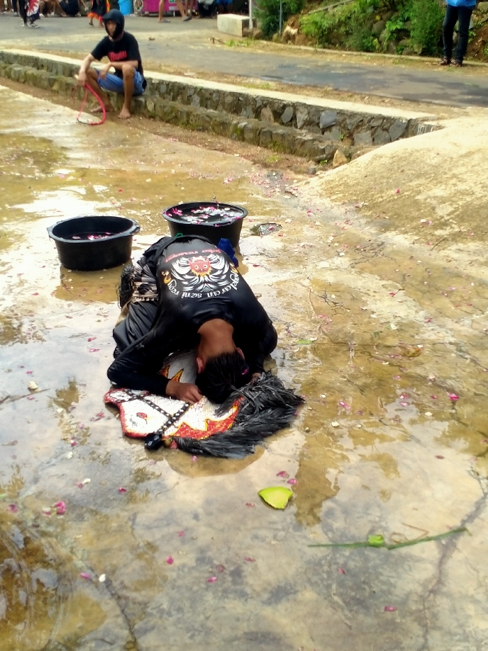 The image depicts a scene on a wet, paved surface in Gebyog, Indonesia, likely outdoors. In the foreground, a person is lying prone, face down, on a concrete ground covered with shallow puddles and scattered pink flower petals. This individual wears a black long-sleeved shirt featuring a prominent white and orange winged emblem on the back, and black trousers. Their head rests on a decorative, stylized horse-head object with white, red, and black patterns and black feathery adornments.
Adjacent to the prone individual, two black plastic tubs contain dark water and additional pink flower petals. In the mid-ground, a male figure sits barefoot on a low, tiered concrete retaining wall. He wears a black hooded top, blue jeans, and holds a flexible reddish-pink loop. Behind this wall, a paved road or path is visible, with green foliage and an earthen bank ascending beyond it. The legs of another person, wearing blue trousers, are partially visible standing near the road on the far right. Indistinct figures are also discernible in the far background on the left side. The ground appears uniformly wet, suggesting either recent precipitation or an intentional use of water.