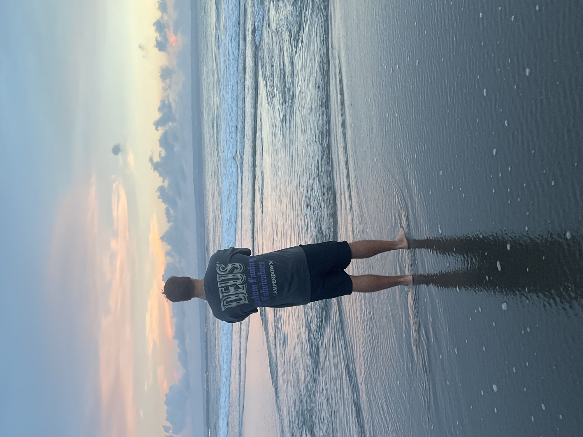 A man stands barefoot at the shoreline of a beach in Kerobokan Kelod, Indonesia, with gentle waves lapping at his feet. He is facing the ocean, his back to the camera, wearing a dark t-shirt with lighter text. The sky is visible above the water, exhibiting soft colors of dusk or dawn. The beach sand is wet and reflects the sky.