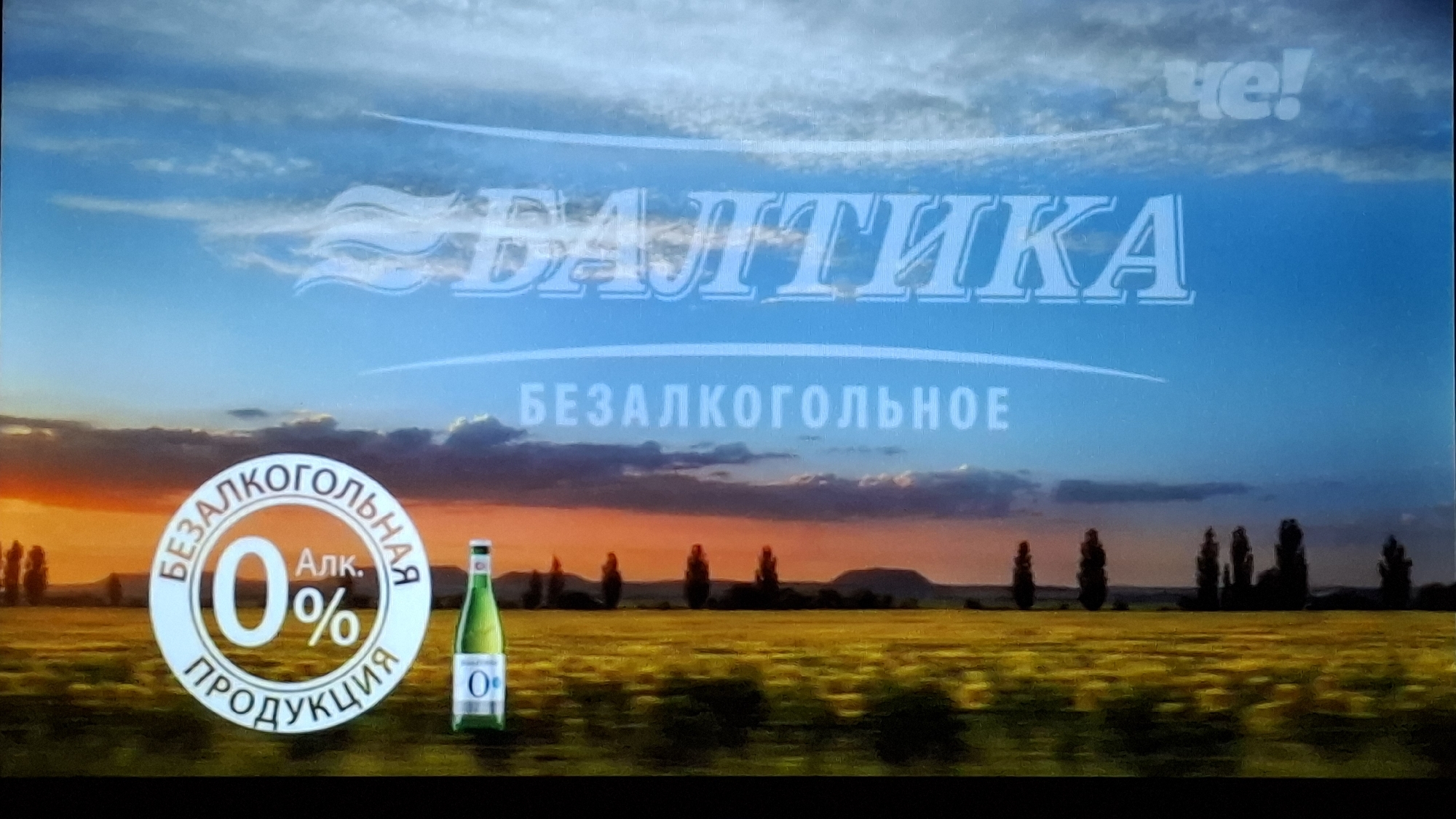 This image is a still frame from an advertisement, likely for a non-alcoholic beverage from the Russian brand Baltika. The scene depicts a serene landscape, likely at sunrise or sunset, featuring a wide expanse of a golden-yellow field, possibly rapeseed or wheat, under a vast sky.
The upper portion of the image is dominated by a clear, bright blue sky with wispy white clouds. The horizon, stretching across the middle of the frame, exhibits warm orange and pink hues, indicative of either dawn or dusk. Along this horizon, a line of silhouetted trees and possibly distant hills adds depth to the rural setting.
Prominently displayed in the upper middle of the sky is the brand name "БАЛТИКА" (BALTIKA) in large, stylized white letters. Below it, the text "БЕЗАЛКОГОЛЬНОЕ" (NON-ALCOHOLIC) explicitly states the product's characteristic. In the top right corner, a white logo for "ЧЕ!" (CHE!), a Russian television channel, is visible.
On the left side of the golden field, a large, circular white and orange emblem highlights the product's key feature: "БЕЗАЛКОГОЛЬНАЯ" (NON-ALCOHOLIC) "0% Алк." (Alcohol 0%) "ПРОДУКЦИЯ" (PRODUCT). Adjacent to this logo, a green-tinted glass bottle, typical of a beer or similar beverage, stands in the field, clearly showing a "0%" mark on its label.
The image conveys a sense of natural beauty and freshness, using the picturesque landscape to promote a non-alcoholic product. The time of day suggests a peaceful and refreshing experience associated with the beverage.