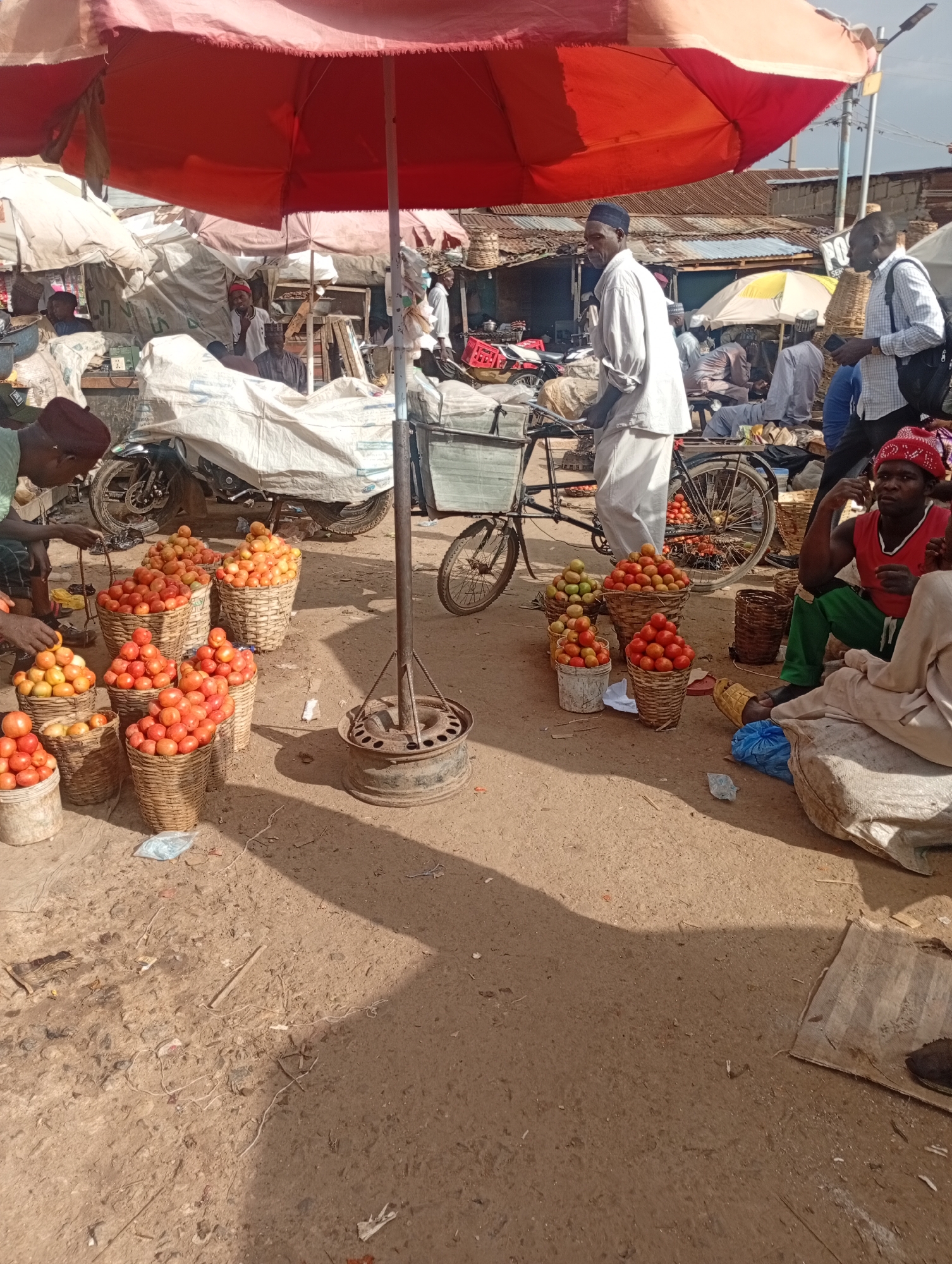 This is a medium shot, eye-level, outdoor photograph of a market scene in Rafin Par, Nigeria. A large, orange and white striped umbrella dominates the upper portion of the frame, providing shade over a market stall. Beneath the umbrella, several people are visible. In the foreground, baskets filled with ripe tomatoes are arranged on the ground. A man in a red sleeveless shirt and a red hat is seated on the right, looking towards the viewer. He is wearing green trousers. To the left, a man in a light-colored shirt and trousers stands and appears to be interacting with the produce. Behind him, a motorcycle is covered with a white tarp. Further back, other market stalls with various goods and more people are visible, suggesting a busy market environment. The ground is dusty and appears to be packed earth. The overall scene depicts a typical day at a Nigerian open-air market.