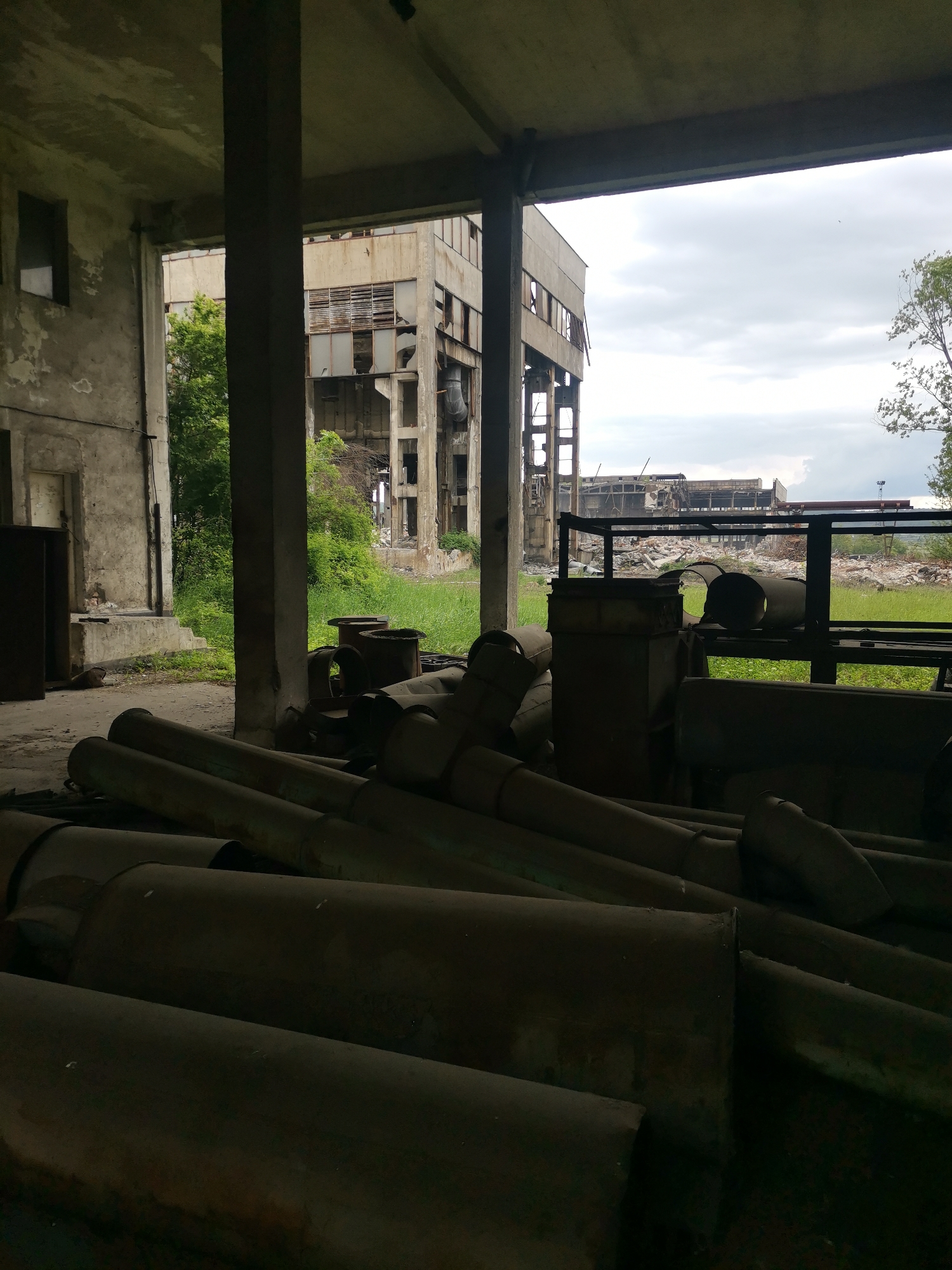 The image depicts the interior of an abandoned industrial structure in Zhivkovo, Bulgaria. Cylindrical metal pipes are piled in the foreground, occupying much of the lower part of the frame. Behind the pipes are metal structures and debris. The interior space is concrete with pillars and openings that frame the view of other dilapidated industrial buildings outside. Vegetation is visible growing around the exterior buildings, indicating a state of disuse and decay. The sky is overcast. No people are visible in the image.