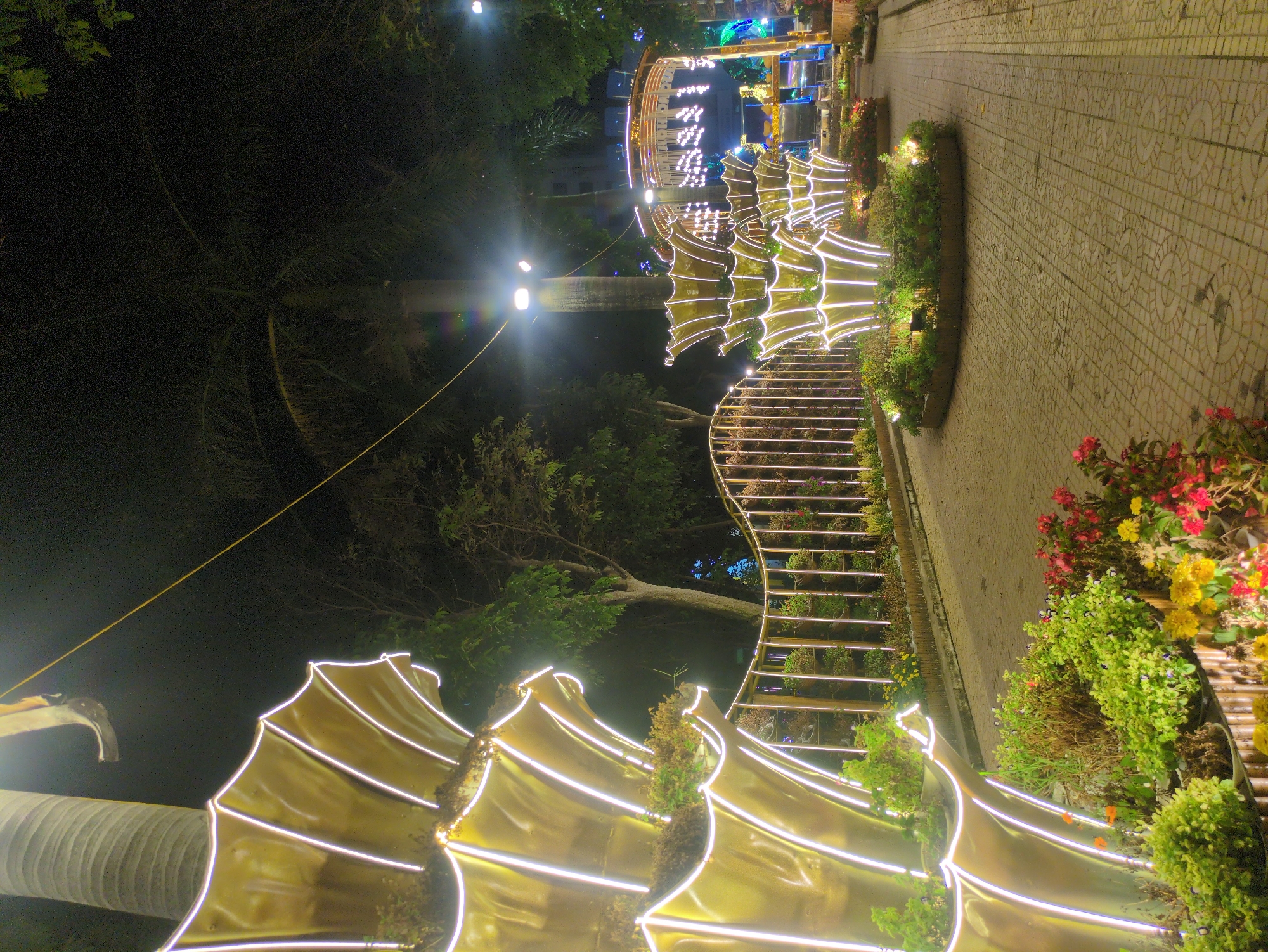 The image captures a night scene in Tuy Hòa, Vietnam, showing a pathway lined with illuminated, decorative structures. The structures appear to be crafted from gold-colored material with integrated white LED strip lights, resembling stylized shells or waves. They are placed along a brick-paved pathway, interspersed with planters filled with greenery and colorful flowers. To the left, a tall palm tree and other foliage contribute to the nighttime atmosphere. In the distance, additional illuminated decorations and structures are visible, adding depth to the scene. The overall lighting creates a warm, inviting ambiance along the path.