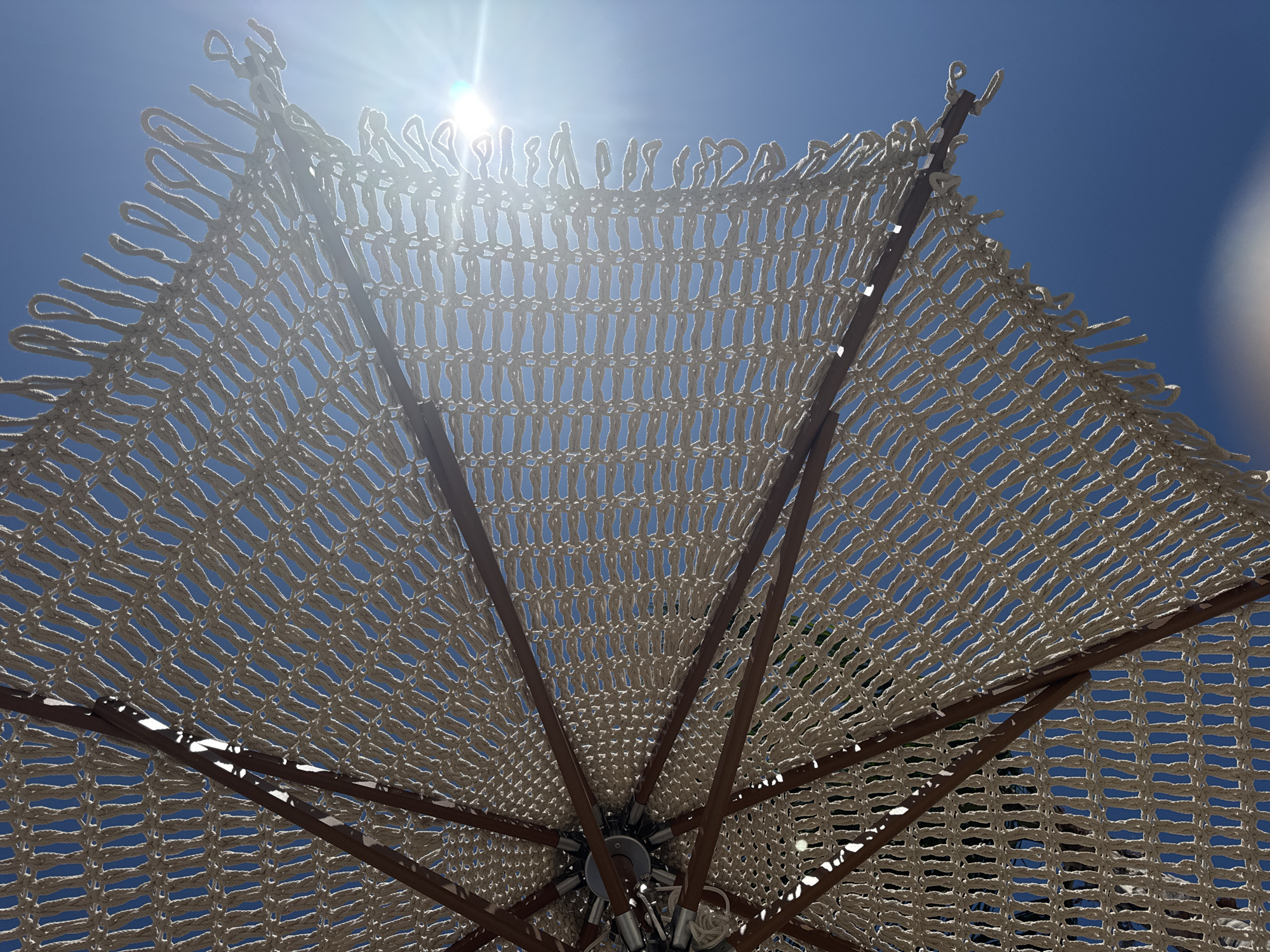 A close-up, low-angle view captures the underside of a white macrame umbrella, revealing its intricate woven pattern against a clear, bright blue sky. The sun, a brilliant white light source, shines from the upper right quadrant, creating lens flare effects and casting a glow that illuminates the netting. Wooden spokes radiate from the center mechanism of the umbrella, supporting the macrame canopy. The overall impression is one of a sunny day, likely outdoors, possibly at a beach or outdoor seating area in Stavros, Greece, given the clear sky and umbrella.