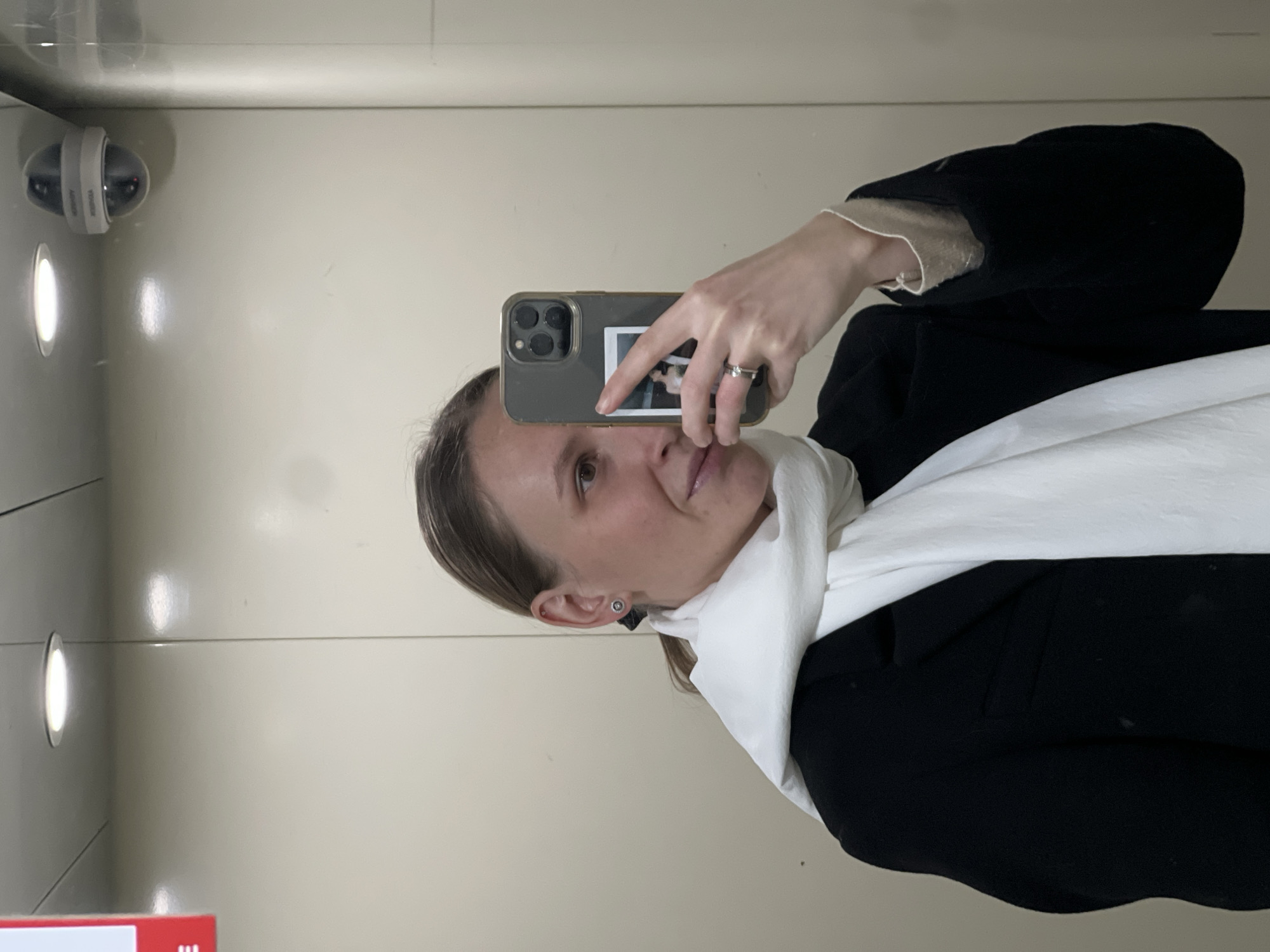 The image shows a woman taking a selfie in what appears to be an elevator. The elevator's interior is white with recessed lights. A security camera is mounted in the upper left corner. The woman is wearing a black blazer and a white scarf. She is holding a smartphone with a phone case and a photograph adhered to it, partially obscuring her face. The photograph suggests a close-up mirror selfie, which is common. The image does not provide any visual information to indicate the location is in Khimki, Russia.