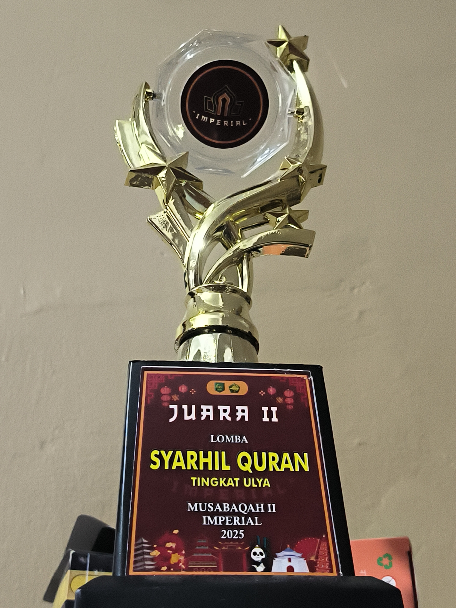 A close-up shot of a trophy with a gold-colored metal body, a clear plastic cup-shaped top with a logo inside, and a black base. The plaque on the base reads "JUARA II" in white text above "LOMBA SYARHIL QURAN" in yellow text, followed by "TINGKAT ULYA" in white. Below that, it reads "MUSABAQAH II IMPERIAL 2025" in white. The plaque's border has a red-brown background with small images of lanterns and other symbols, and at the bottom, there are images of Asian architecture and a panda figurine. The background is a plain beige wall. Lhokseumawe, Indonesia, is not depicted in this image.