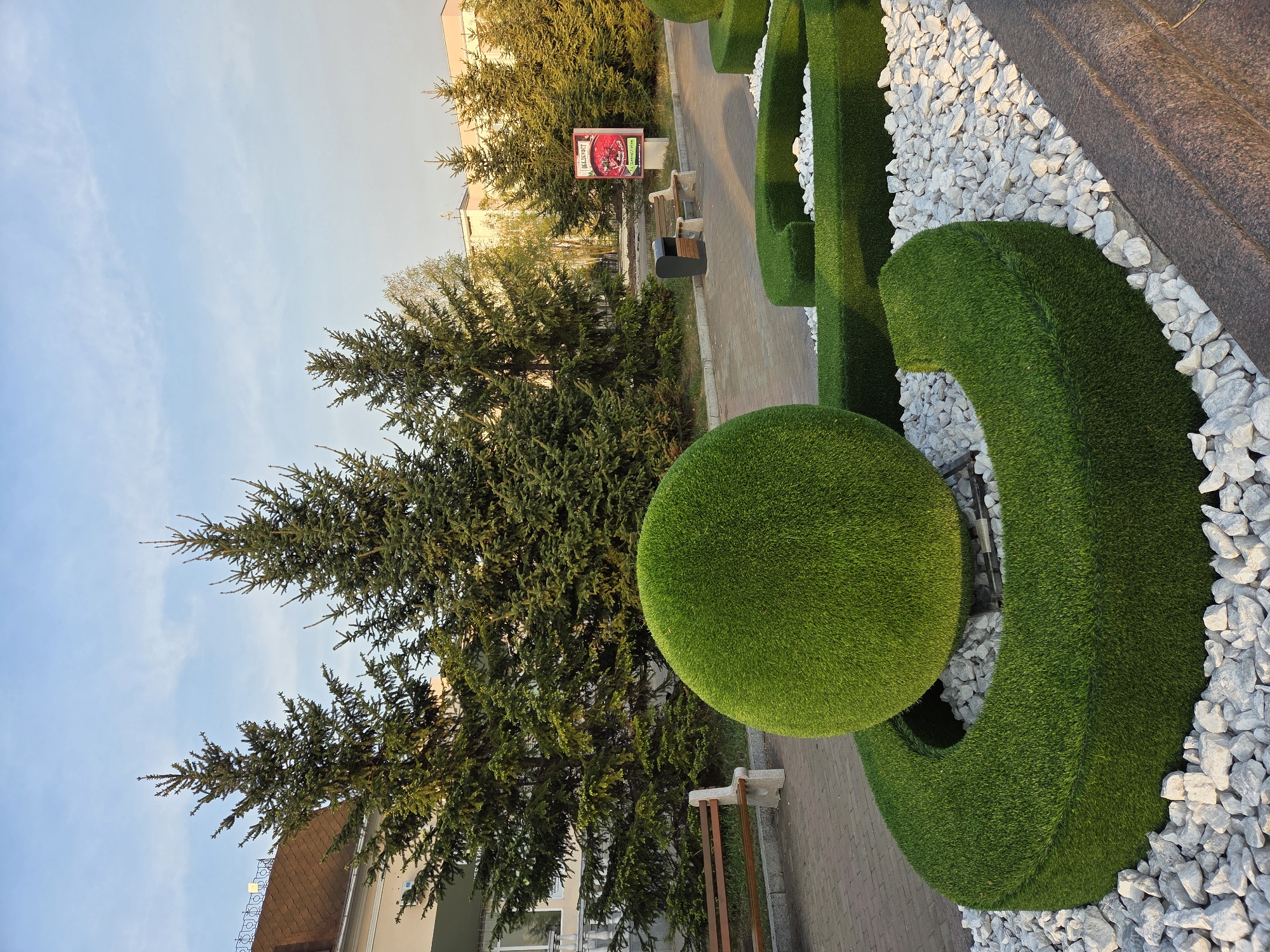 The image shows an outdoor scene in Birobidzhan, Russia, with a large artificial green telephone sculpture made of grass or turf as the focal point. The sculpture is placed on a bed of white rocks or pebbles. A paved walkway is visible next to the sculpture, with a bench nearby. In the background, there are trees, including evergreen trees. Buildings can be seen in the background as well. The sky is visible in the upper portion of the image. There is a sign or advertisement visible near the bench in the background.