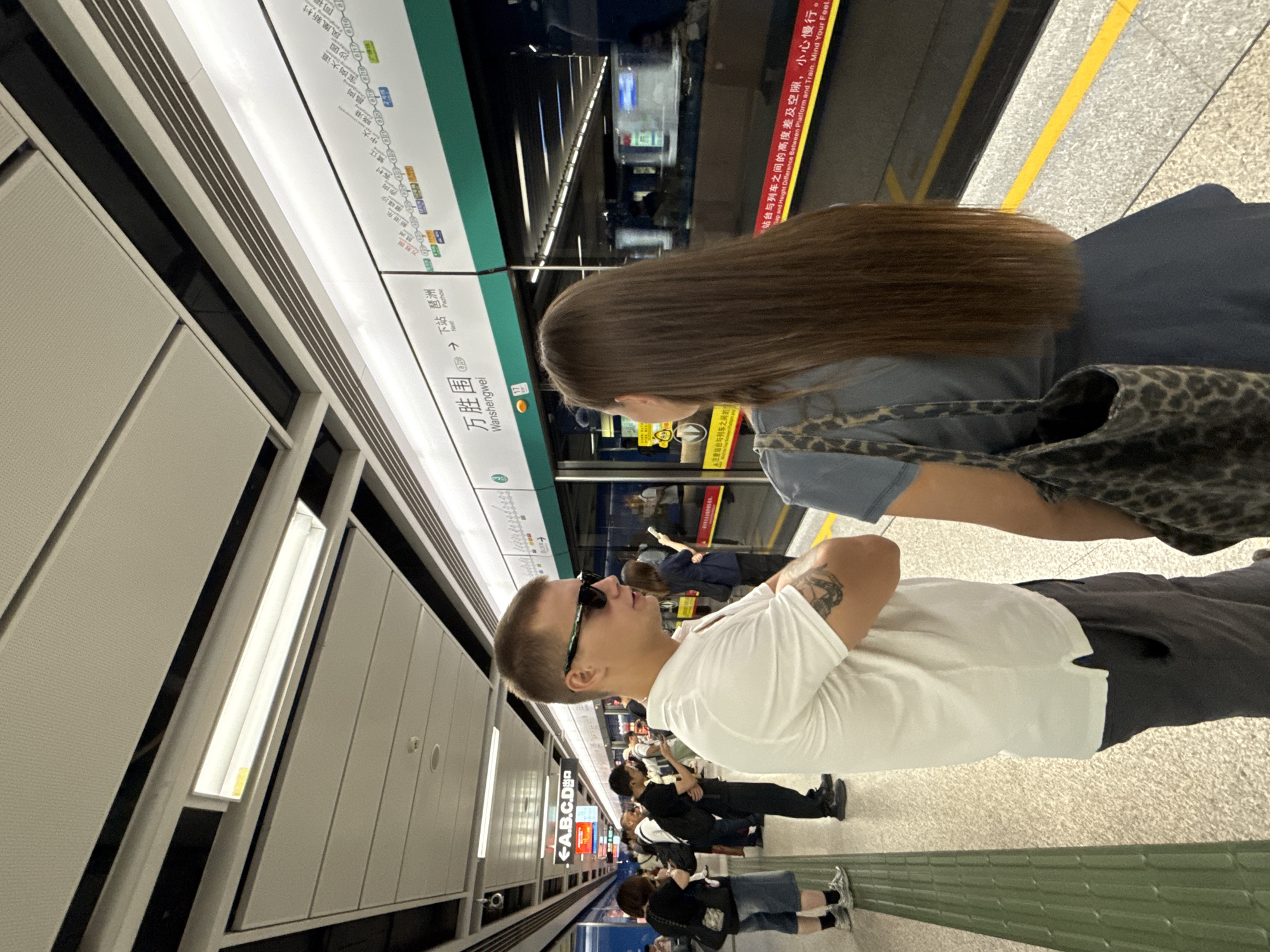 The image shows a man and a woman standing on a subway platform in Haizhu District, China. The man is wearing a white polo shirt and sunglasses. The woman is wearing a blue dress and carrying a leopard-print bag. They are both looking at a subway train that is stopped at the station. The platform is clean and modern, with a map of the subway lines displayed above the train. The lighting is bright, and the overall atmosphere is one of a typical urban subway station.