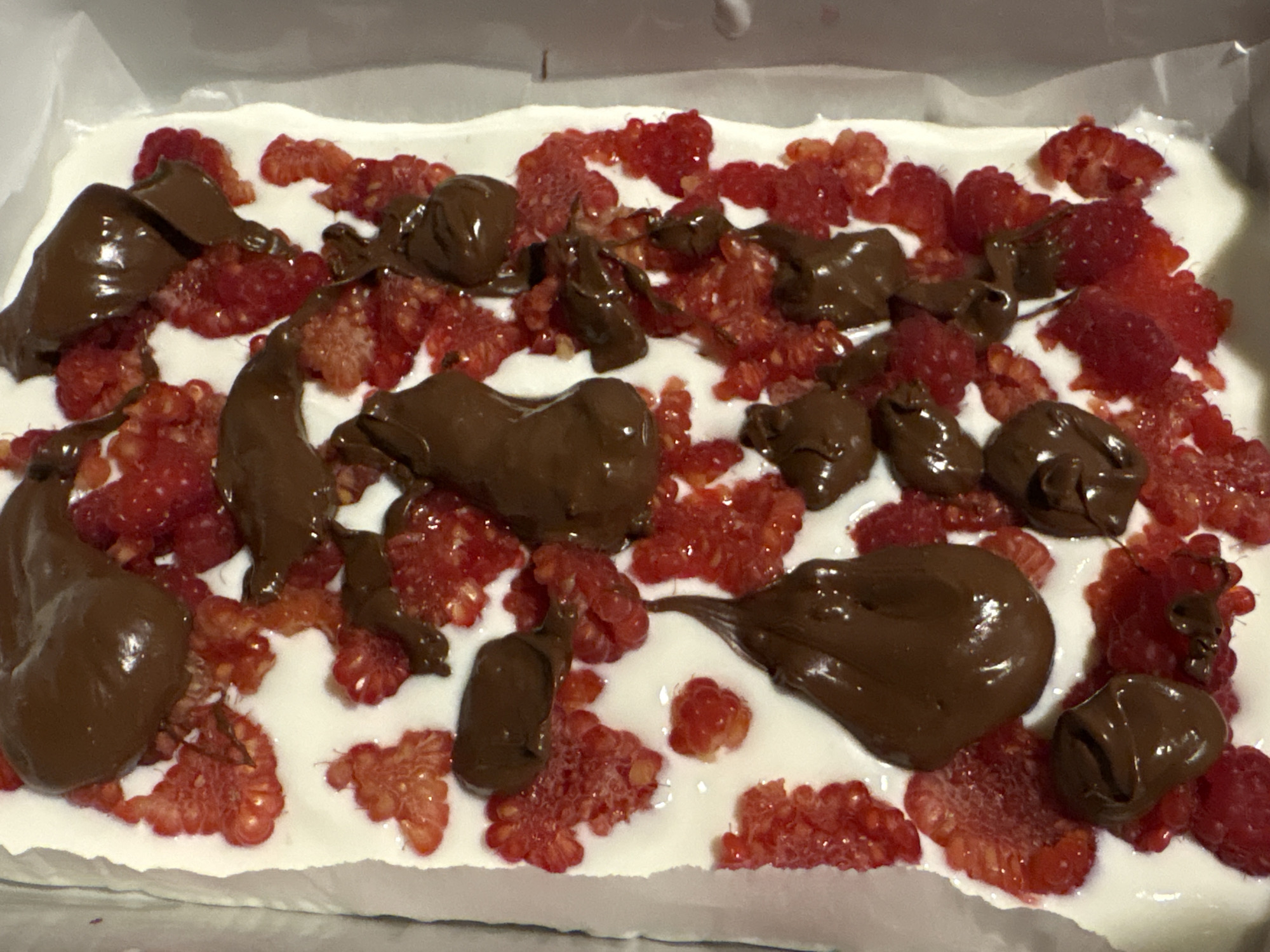 A close-up shot depicts a dessert item, seemingly prepared in a rectangular dish lined with what appears to be parchment paper. The base layer is a white, creamy substance. This base is covered with a layer of red raspberries, and dollops of dark brown chocolate are scattered throughout. The dessert is situated indoors, but no further details about the location are discernible.
