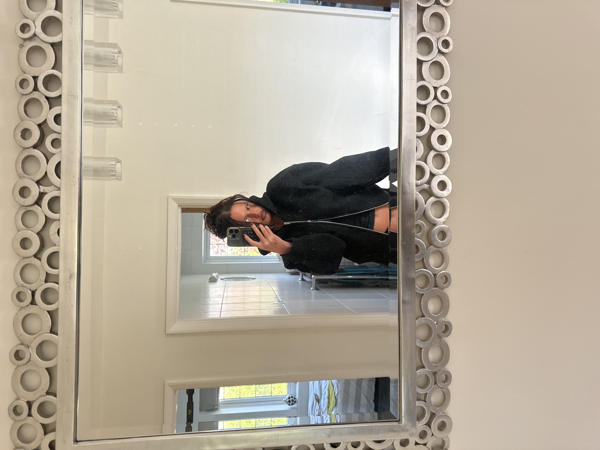 The image depicts a mirror reflection of a person in what appears to be a bathroom. The mirror has a decorative frame composed of circular elements. The person is holding a smartphone, presumably taking the photograph. The reflection reveals a bathroom interior with white walls, tiled floors, and visible fixtures like a shower and a window. There is also a glimpse of a window with a view outside, revealing greenery, suggesting an outdoor environment. The context of Tunbridge Wells, United Kingdom, is based on the provided location.
