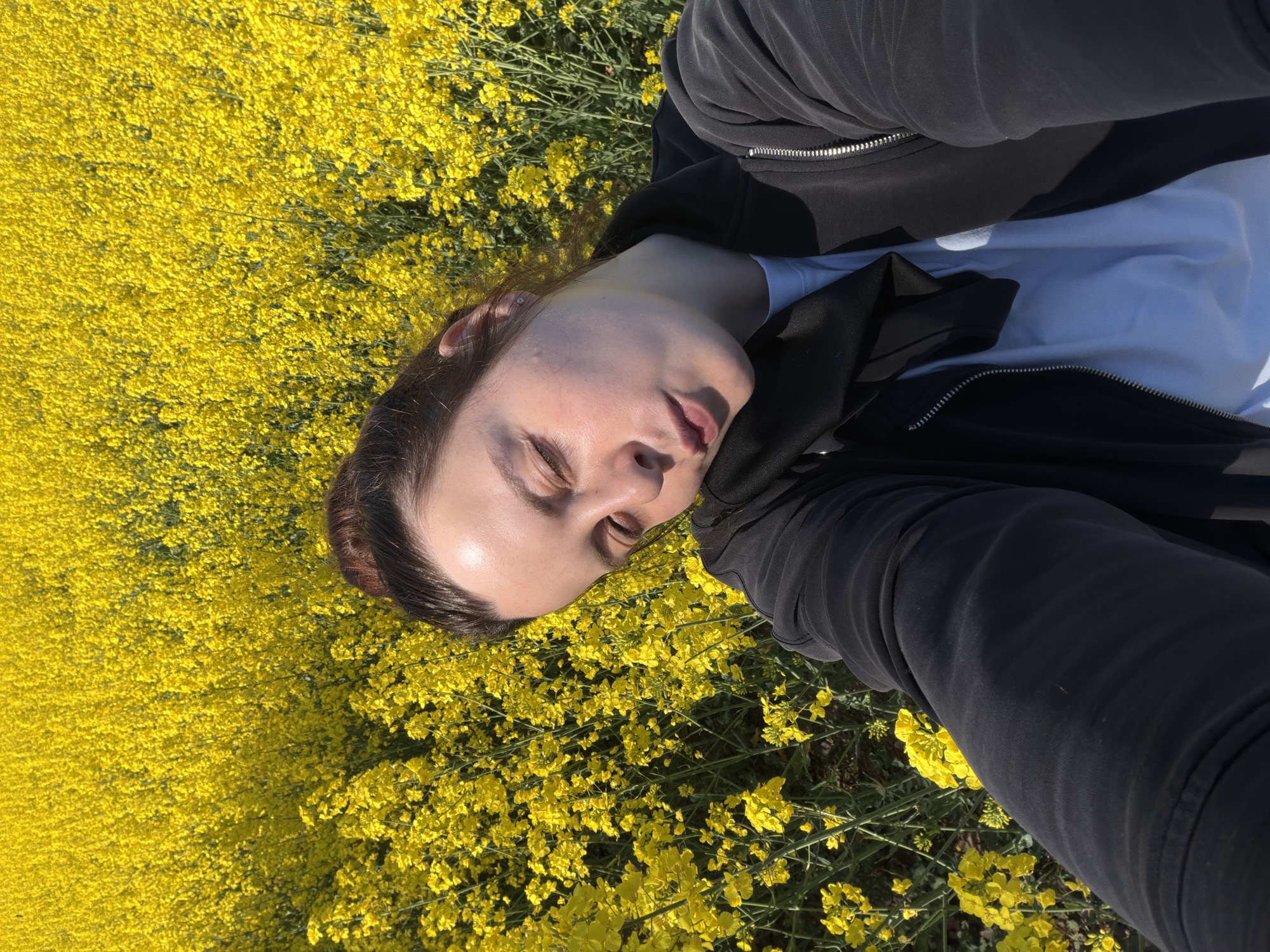 An individual is captured in a top-down selfie, lying amidst a vibrant field of yellow flowers, likely canola or rapeseed, under a bright, sunny sky in Obermoschel, Germany. The person is dressed in a black hooded jacket or sweatshirt worn over a light blue t-shirt. Their hair is dark brown, and their eyes are mostly closed or squinting, suggesting relaxation or an effort to shield from the bright sunlight. The light casts distinct shadows, indicating a clear, sunny day, likely during the morning or afternoon. The dense, brilliant yellow blooms dominate the background, creating a striking contrast with the person's attire and skin tone. No text is visible in the image. The overall mood appears peaceful and serene, with the person enjoying the natural setting.