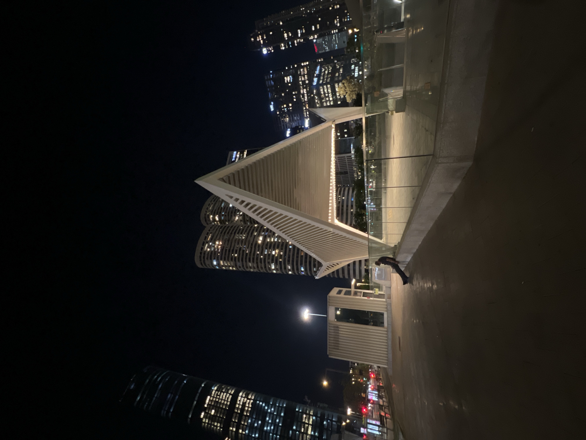 The image shows a nighttime scene in Tel Aviv, Israel. A triangular architectural structure with white beams is centrally positioned, with illuminated skyscrapers rising behind it. In the foreground, a person is bent over near a glass barrier. A white rectangular building with windows is also visible, along with streetlights and blurred traffic signals indicating a street or road below.