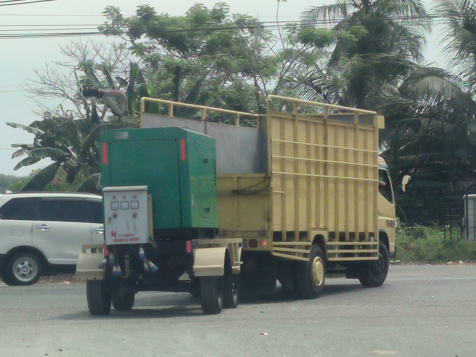 A medium shot of a yellow truck with a green generator mounted to the back driving on a gray asphalt road. The generator has an attached grey electrical panel with the words "PT KESMA PERDANDHA ENERGY". Behind the truck to the left is a white minivan with tinted windows. Lush green foliage is in the background, including banana trees and palm trees. Overhead power lines are visible. The scene is likely in the city of Lhokseumawe, Indonesia, based on the text on the electrical panel.