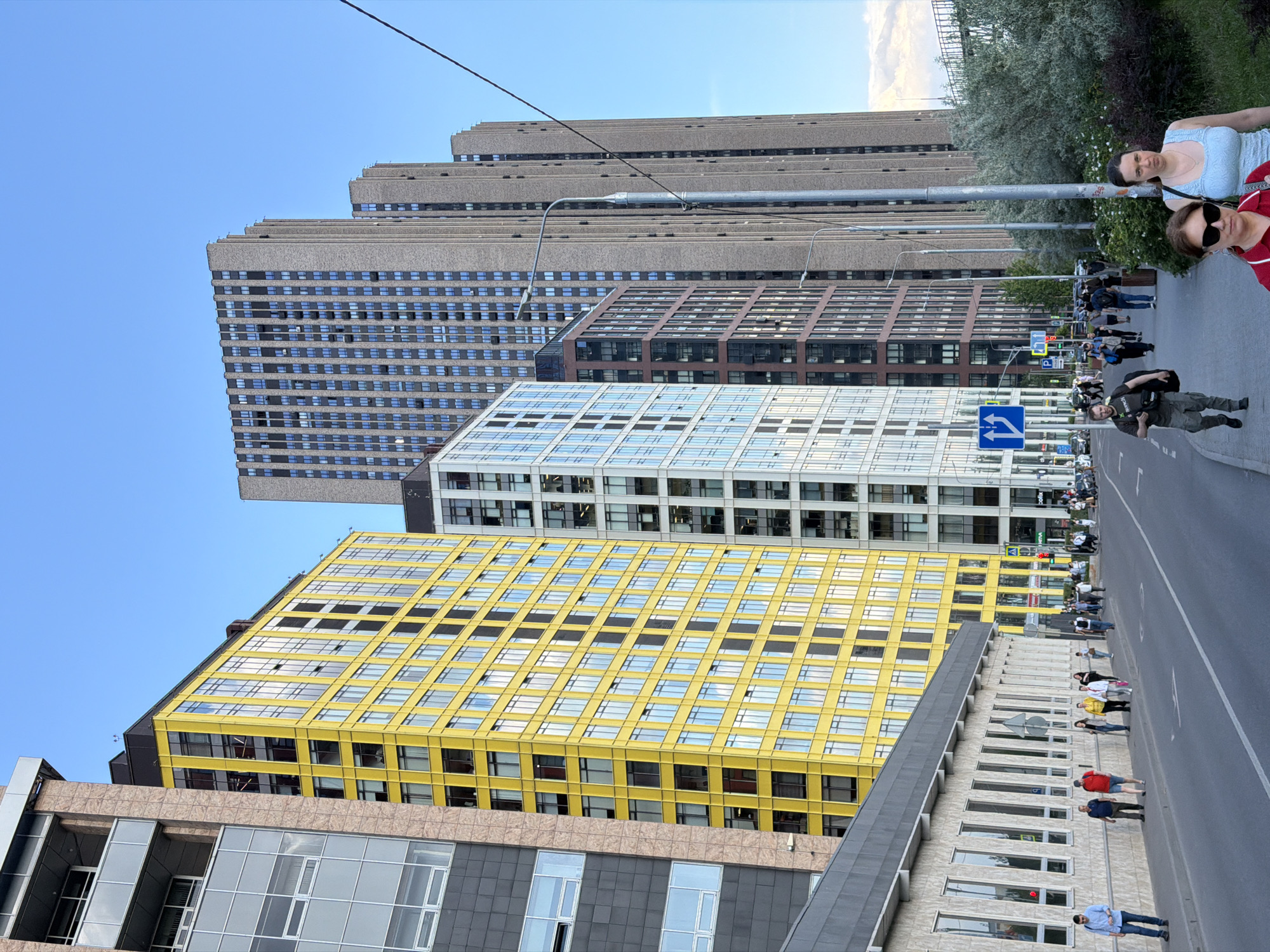 This media file captures a sunny urban street scene in Khimki, Russia, featuring a mix of modern architectural styles. The view looks down a street lined with several prominent buildings under a clear blue sky.
In the background, a cluster of tall, stepped high-rises with grey concrete facades and numerous small, dark windows dominates the skyline. Below these, a building with a reddish-brown brick-like exterior is visible. Further forward, a white building characterized by a grid of large windows and white panels stands out. Adjacent to it, a bright yellow building with a distinct grid pattern of reflective windows adds a bold splash of color to the urban landscape. In the lower right foreground, a building with a dark grey sloped roof and tall, narrow windows is partially visible.
The street is paved with asphalt and features a white dashed line. Pedestrians are scattered along both sidewalks and crosswalks. On the right, closer to the viewer, a woman in a light blue top and another in a red top wearing sunglasses are visible. On the opposite side of the street, a man in dark clothing, carrying a backpack, walks along. Numerous other people are seen walking further down the street, indicating an active urban environment. Trees and shrubs line parts of the sidewalks, adding greenery to the scene.
A blue directional road sign with two white arrows—one pointing left and the other straight ahead—is visible above the street. Overhead wires stretch across the sky, connected to utility poles. The strong, clear light suggests it is daytime, possibly late afternoon or early evening. No other visible text is clearly discernible.