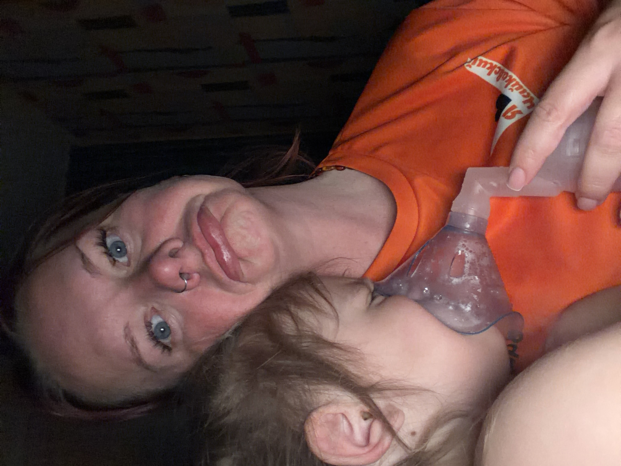A photograph taken indoors depicts an adult woman holding an oxygen mask to the face of a young child. The woman has blue eyes, fair skin, and a nose ring and is wearing an orange t-shirt with white lettering. She is making a funny face with her tongue sticking out. The child has light brown hair and fair skin, and is lying with their head against the woman’s chest. The room is dimly lit. The location is Unknown, Unknown.
