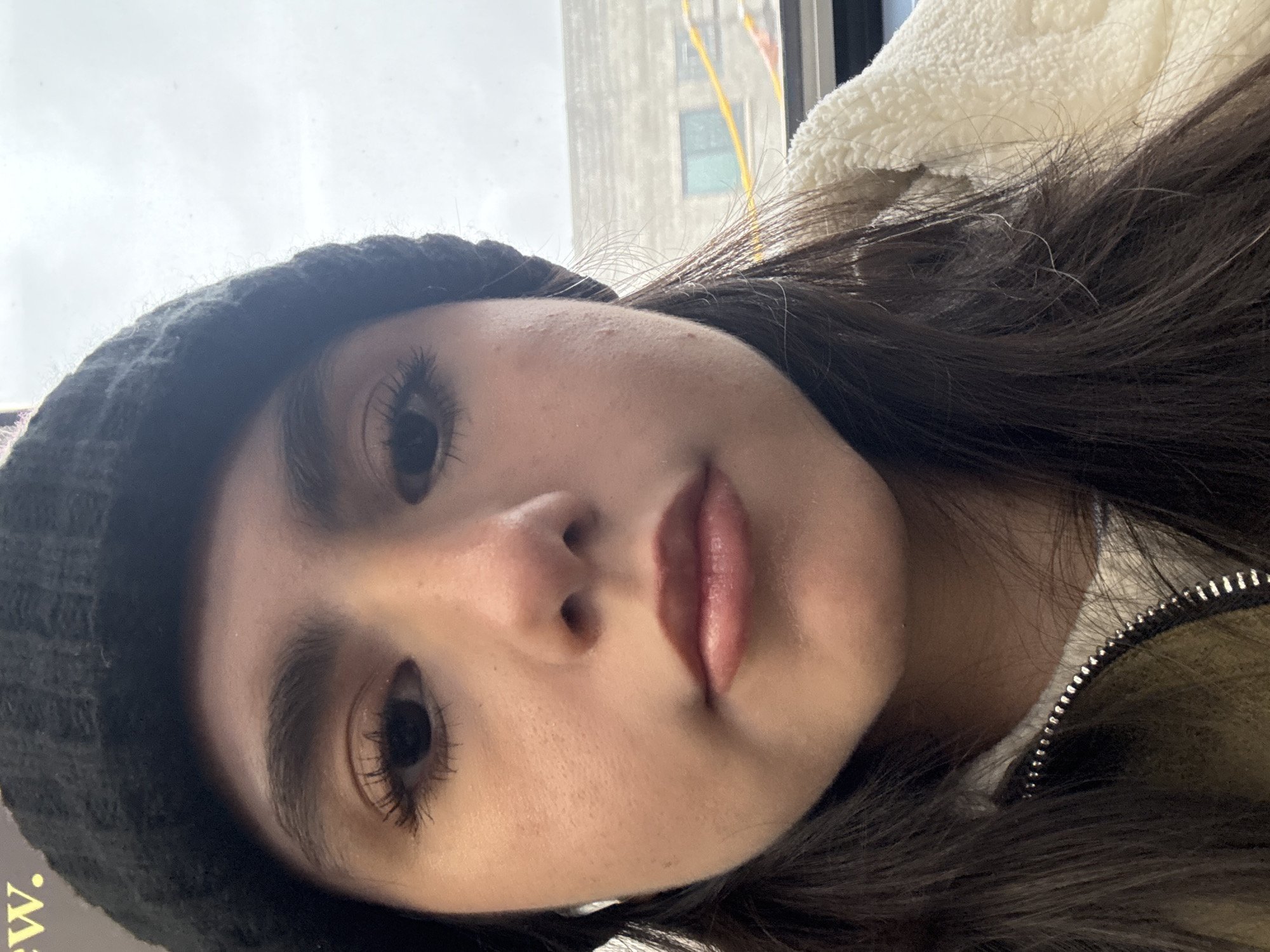 A close-up, rotated selfie shot depicts a young woman wearing a black knit cap. She has dark hair, long eyelashes, and full lips. She is wearing a white, textured jacket with a visible zipper. The background shows a city building visible through a window, consistent with Chicago architecture. Given the urban backdrop, the woman is likely inside a vehicle or building with a view of the city.