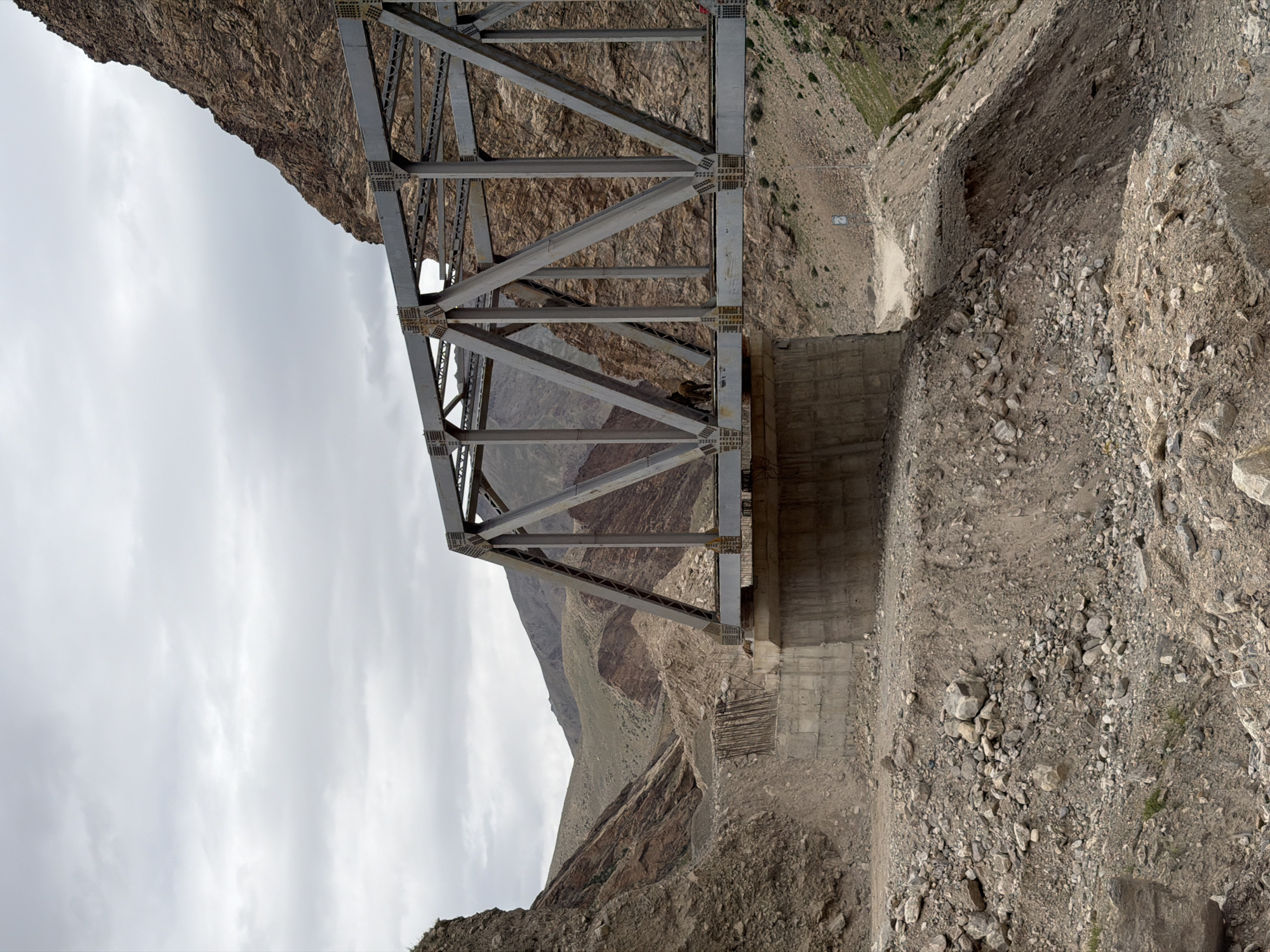 The image depicts a steel truss bridge spanning a rocky, mountainous terrain. The bridge is gray and constructed with a lattice of diagonal beams. It crosses over a dry riverbed or ravine. Concrete abutments support the bridge. The surrounding landscape is characterized by steep, barren slopes covered in loose rocks and gravel. Vegetation is sparse, with a few patches of dry grass and scrub. The sky is overcast with gray clouds. A dirt road winds its way up the mountainside on the right side of the image. There is a single person, appearing to be a male, standing on the bridge, looking away from the camera. Another person is partially visible on the far right of the bridge structure. The scene appears to be in a remote, undeveloped area. The location is identified as Reru, India.