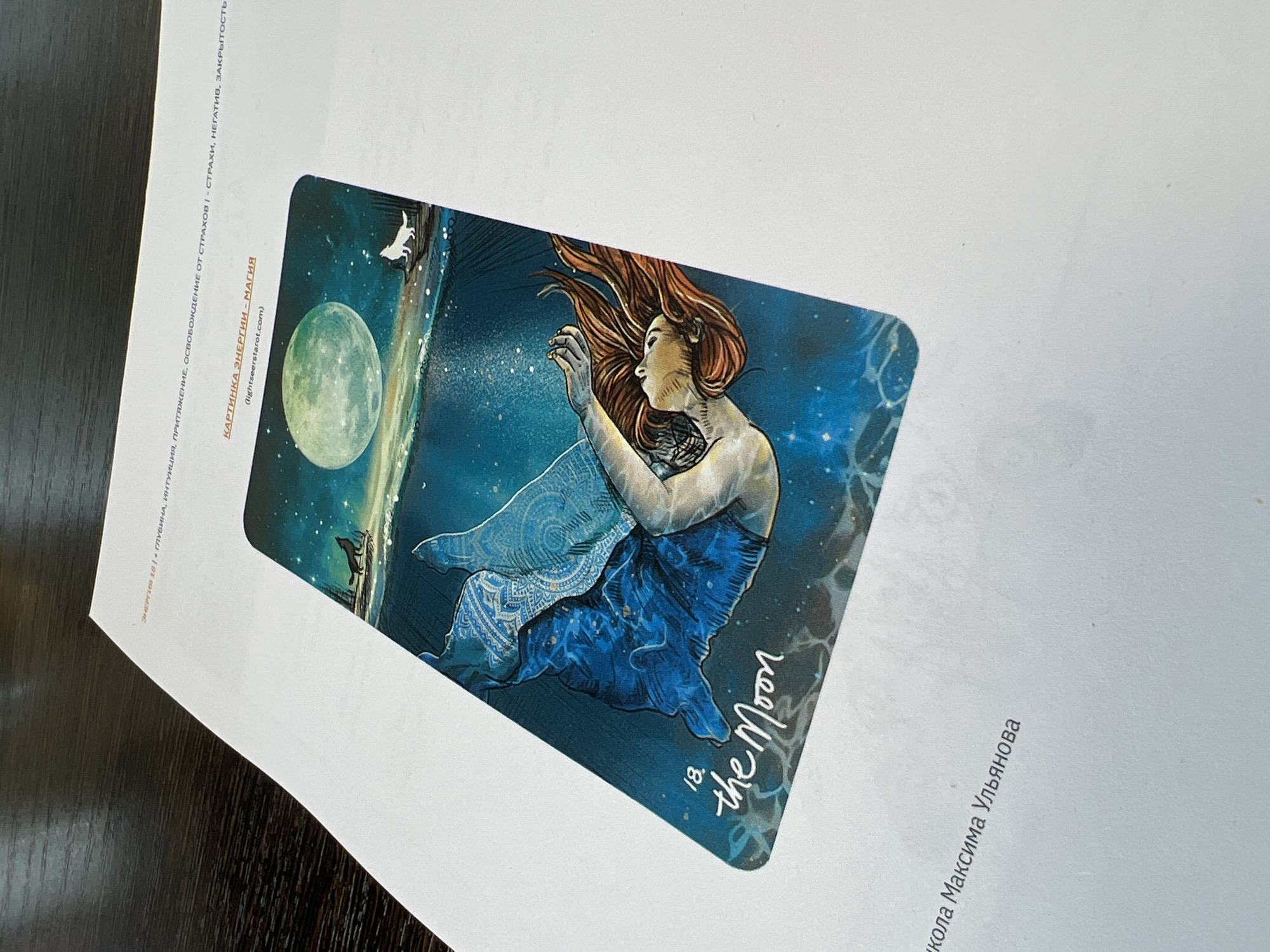 The image shows a Tarot card with the number 18, titled "The Moon." It depicts a woman with flowing red hair in a blue dress. The background is a night scene with a full moon, stars, a wolf, and another animal, possibly a dog. The text on the printed paper next to the card includes Russian phrases related to energy, intuition, attraction, and freedom from fears. Below the card are the words "Никола Максима Ульянова" and "КАРТИНКА ЭНЕРГИИ - МАГИЯ (lightseerstarot.com)." The location is unknown and nothing in the image indicates it is in Perm, Russia.