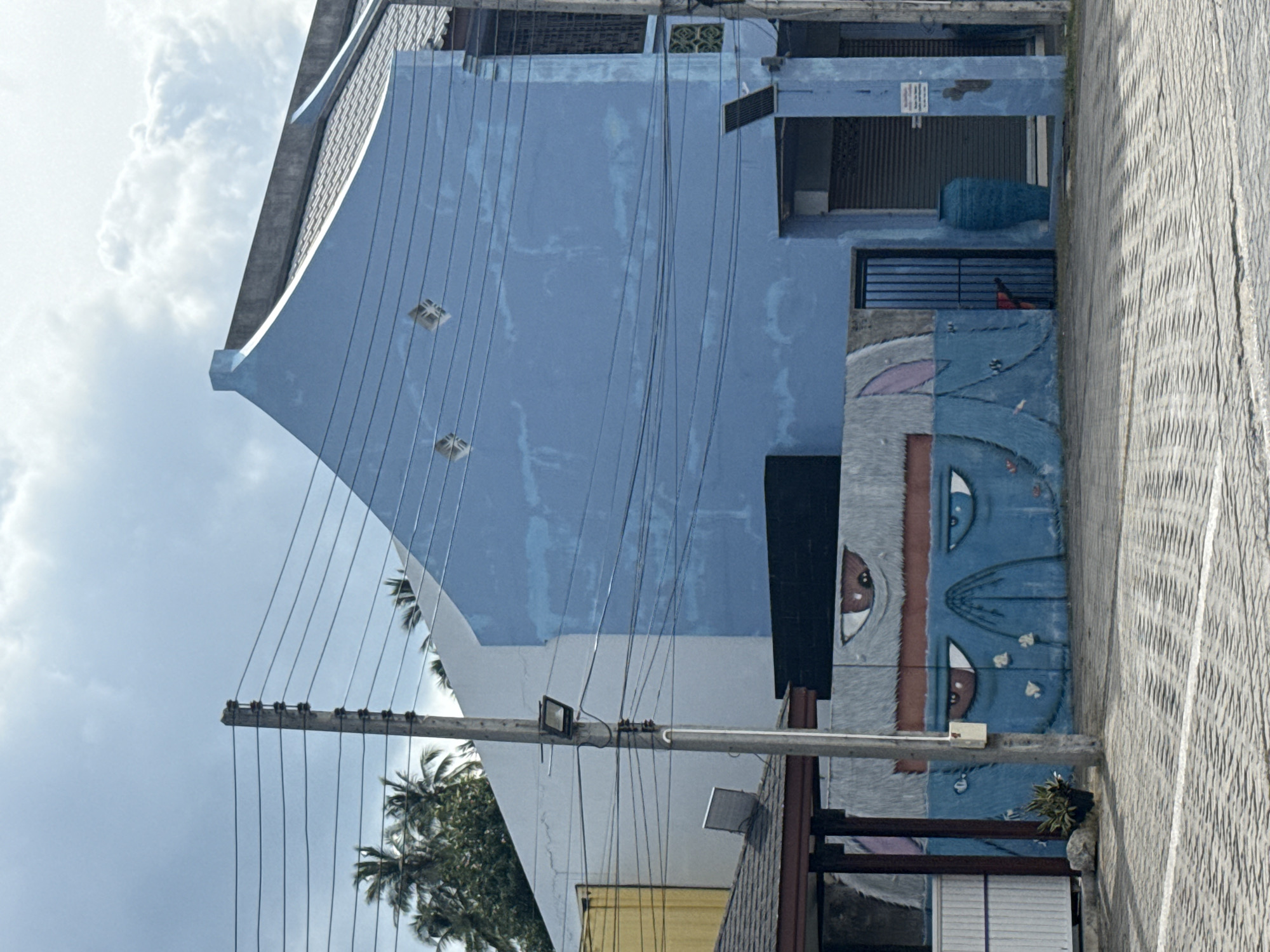 The image depicts a street scene in the city of Ko Samui District, Thailand, under a cloudy sky. In the foreground, a paved walkway with a white line runs horizontally. A concrete utility pole with multiple wires extending from it stands prominently on the left side of the image. Behind the pole, a building with a light blue facade is visible, featuring a mural of cartoonish eyes and a furry texture. This building has a dark-colored roof and a small, gated entrance. To the left of the blue building, a white building is partially visible, and further left, a building with a yellow facade can be seen. In the background, palm trees are discernible, suggesting a tropical location. The overall scene appears to be an outdoor area, possibly a street or a plaza, with a mixture of residential or commercial buildings and utility infrastructure.