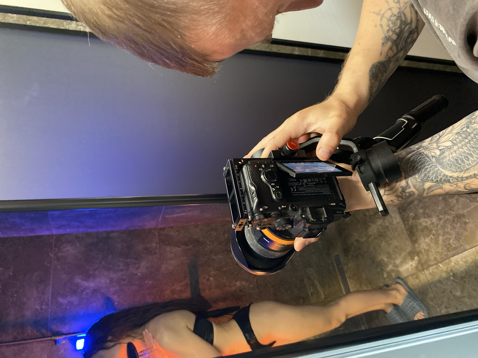 A person is taking a photo of a reflection in a mirror. The reflection shows a person lying on the floor wearing a black bikini. The person taking the photo is holding a professional camera with a large lens and has tattoos on their arm. The environment is modern. There is no indication that the location is Votkinsk, Russia.