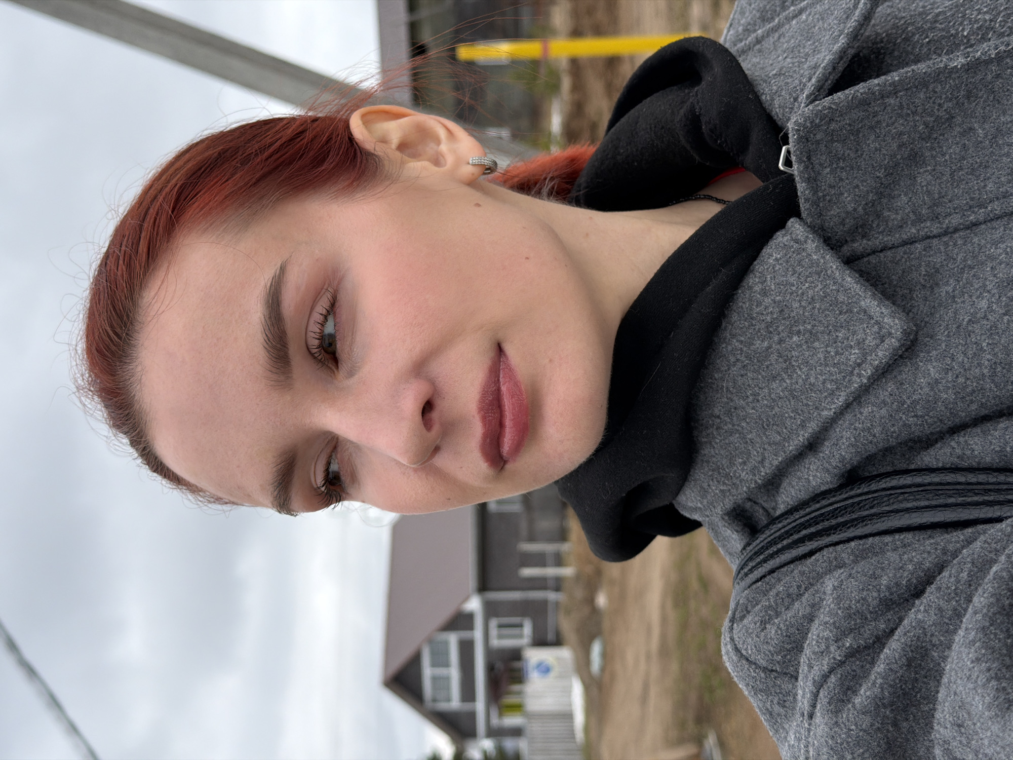 A red-haired woman wearing a gray coat over a black hoodie is shown in an outdoor, close-up shot, with her face angled downward and to the left. The background reveals a cloudy sky and a partially visible house, suggesting an urban or suburban setting. Based on the weather and style, it appears to be taken in Chaikovsky, Russia.