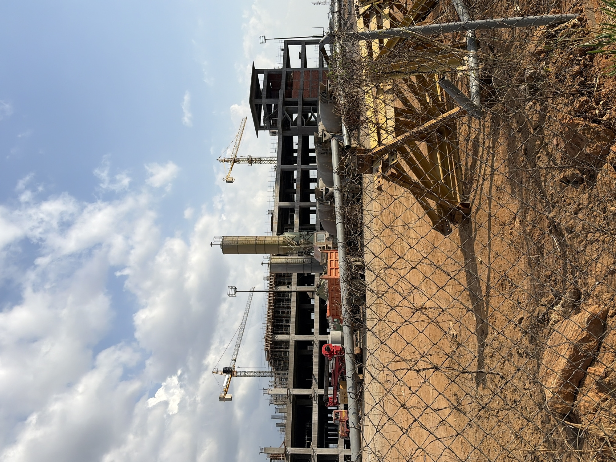 The image shows a construction site in Ciudad Guayana, Venezuela. A metal chain-link fence runs along the foreground, partially obscuring the view of the construction area. Behind the fence, a multi-story building is under construction with a steel frame and some brickwork. Several yellow cranes are visible in the background, indicating active construction work. There are stacks of building materials, pipes, and equipment visible inside the fenced perimeter. The ground near the fence is dirt and soil. The sky is blue with scattered clouds.