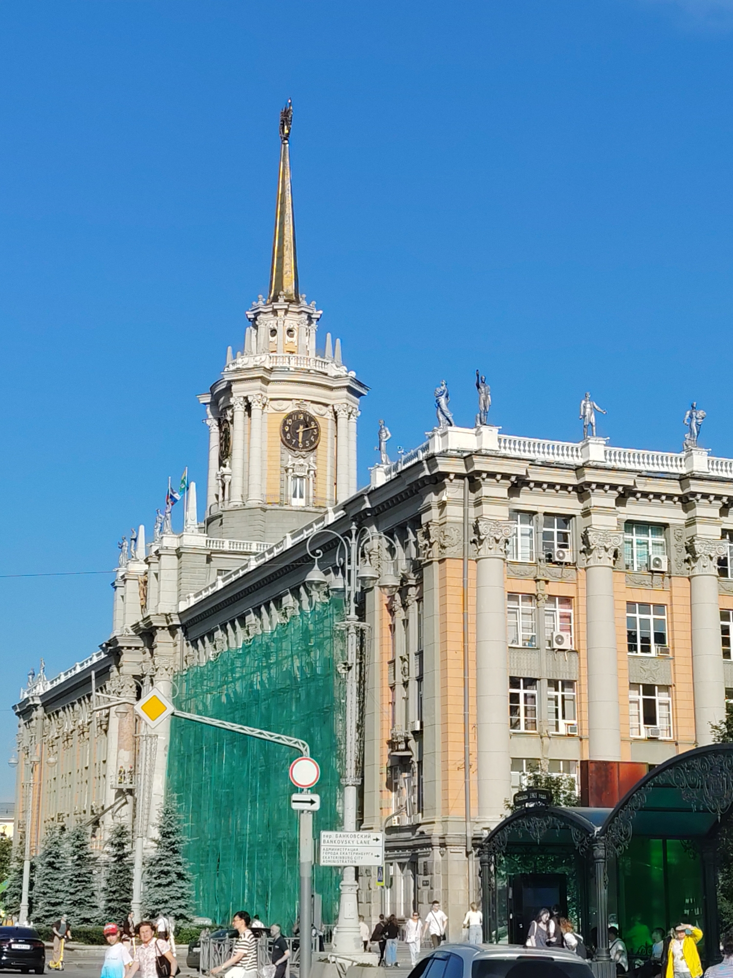 This media file is a photograph of a grand, multi-story building with classical architectural elements, set against a clear blue sky. The building features a prominent central tower topped with a golden spire, adorned with a statue. The tower also has a large clock face. Numerous statues of human figures are placed along the roofline of the building.
The building appears to be undergoing renovation, as a large section of its facade is covered with green netting supported by scaffolding. In front of the building, there is a street scene with several people walking and some standing. A black car is visible on the left side of the image, and a few trees are planted along the sidewalk. There is a distinctive green, ornate glass structure, possibly an entrance or bus shelter, in the lower right corner of the frame. Traffic signs, including a diamond-shaped warning sign and a circular prohibition sign with an arrow, are also visible.
The lighting suggests it is daytime, likely morning or afternoon, with bright sunlight casting shadows. The clear blue sky indicates good weather conditions.
Visible text on a white sign post includes: "ДН. БАНКОВСКИЙ ЛАНЕ" and below it in English: "BANKOVSKY LANE". Further text includes "АДМИНИСТРАЦИЯ" and "YEKATERINBURG CITY ADMINISTRATION".
The overall scene depicts an urban environment in Yekaterinburg, Russia, with a focus on the impressive architecture of the building and the ongoing renovations.