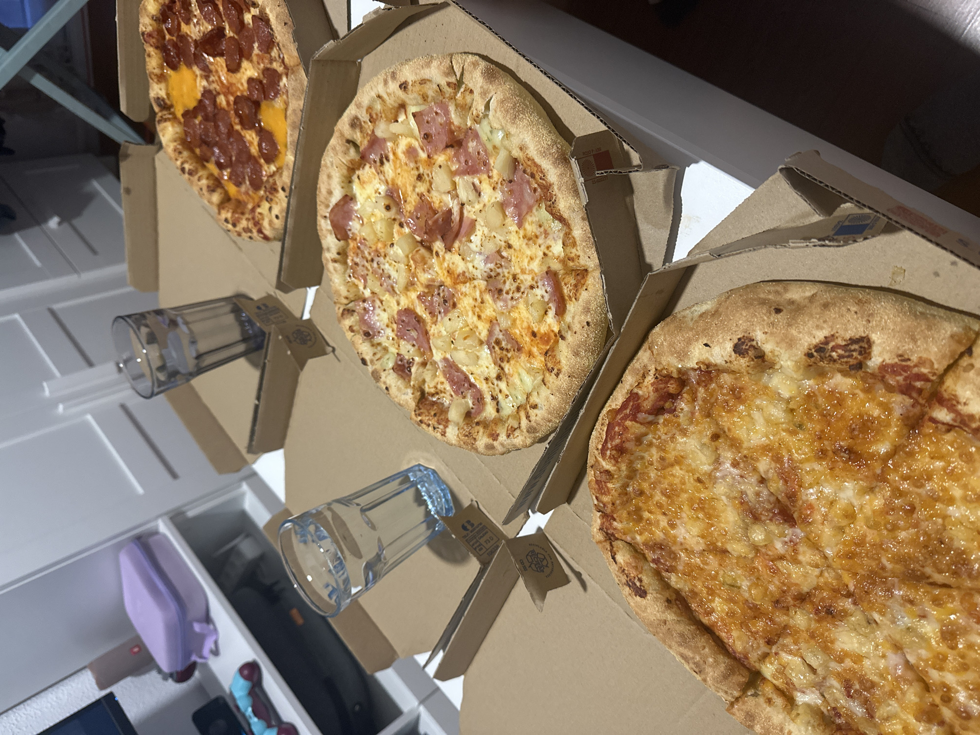 In a brightly lit indoor setting, possibly a home, four pizzas are arranged in cardboard boxes on a white table. The pizzas vary in toppings: one appears to be a classic margherita, another is topped with pepperoni and cheese, a third features ham and pineapple, and the fourth is a simple cheese pizza. Two glasses of water are also present, one on the table and one placed beside a pizza box. In the background, a white shelving unit holds a purple lunchbox and what looks like a video game controller. The scene suggests a casual meal or gathering. The location in Oviedo/Uviéu, Spain, cannot be determined from the visual information provided.