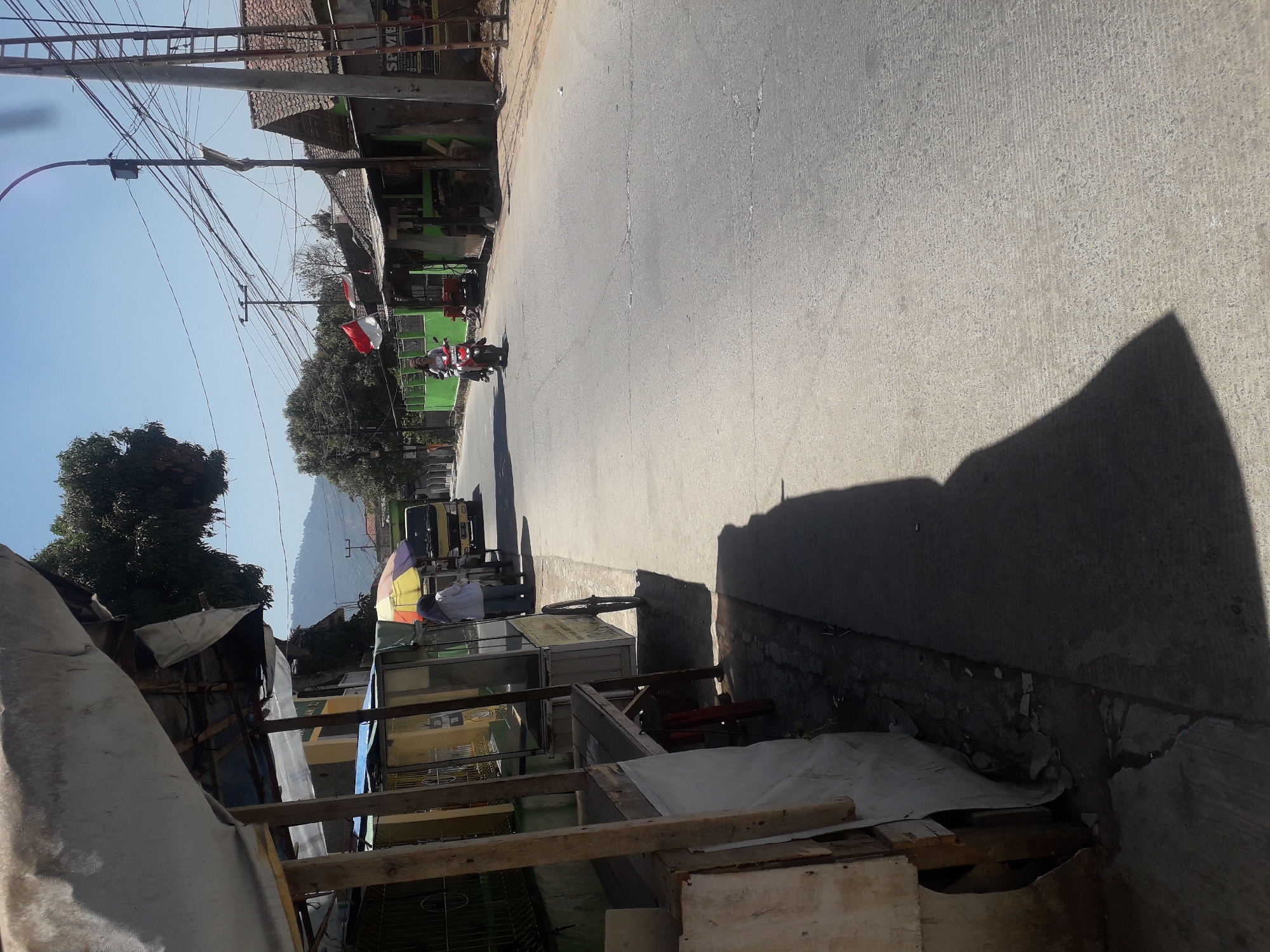 This is a street scene in Cankuang, Indonesia. A person is riding a motorcycle on the right side of the road, heading away from the camera. The road is paved and appears to be a typical village road. On the left side of the road, there is a makeshift stall or shop covered with a white tarp. Behind the stall, there are some structures and more trees. Further down the road on the right, there are several houses with tiled roofs and utility poles with many wires. A small Indonesian flag is visible on one of the houses. The sky is clear and blue, suggesting a sunny day. The overall impression is of a rural or semi-rural environment in Indonesia.