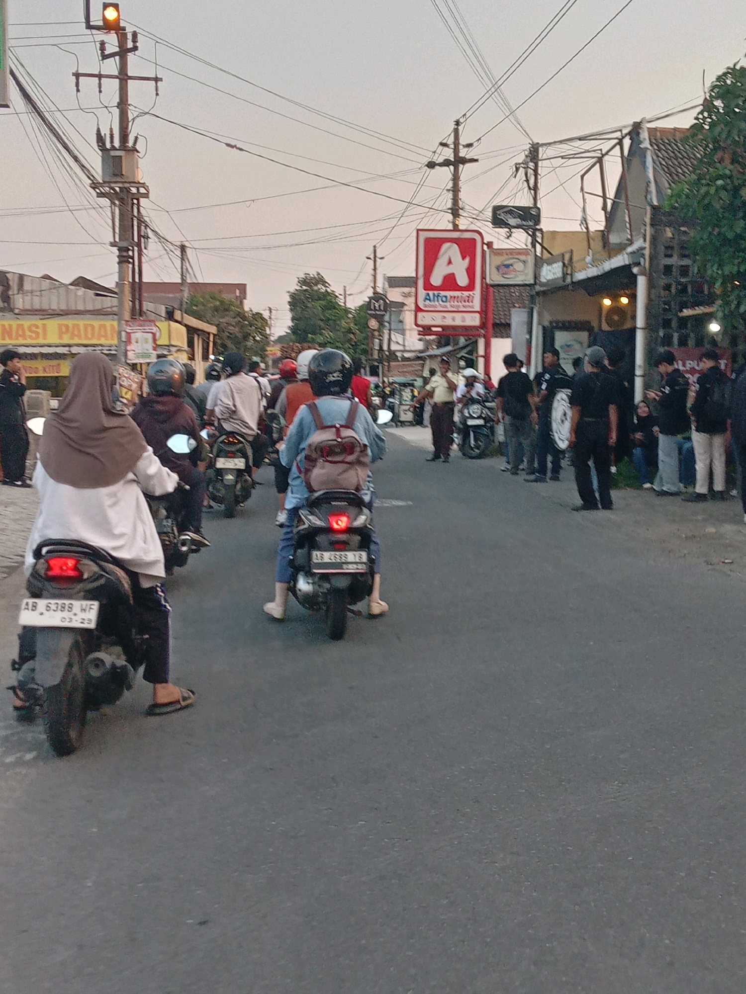 The image depicts a street scene in Banguntapan, Indonesia, likely during the late afternoon or early evening based on the lighting. A traffic light with a yellow signal is visible atop a utility pole, surrounded by a network of overhead electrical wires. Several people are riding motorcycles on the asphalt road. One person in the foreground, wearing a brown hijab and a white outer garment, is on a motorcycle with a license plate reading "AB_6388_WF". Another person in front of them, also on a motorcycle, wears a blue jacket and a backpack. A red sign with a white "A" and the text "Alfamart" is prominent on the right side of the street, indicating a retail establishment. Several other individuals are standing or sitting on the roadside, some wearing black clothing. Buildings with tiled roofs line the street, and trees are visible in the background. The overall atmosphere appears to be that of a busy urban thoroughfare.