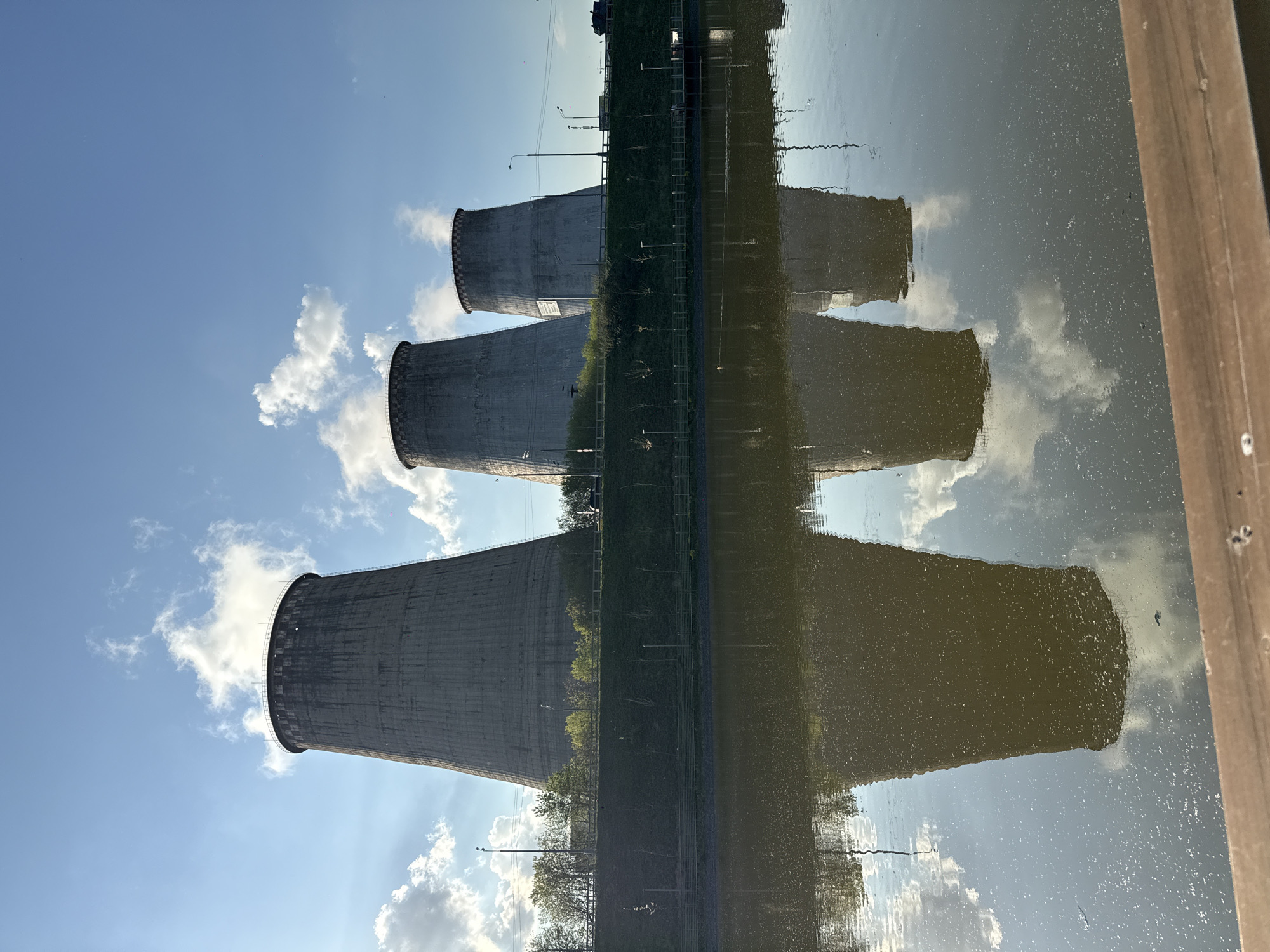 The image depicts three cooling towers reflected in water, likely a reservoir or pond. The towers are made of gray concrete and are arranged vertically in the frame. The sky is blue with scattered white clouds. Between the towers and their reflections is a strip of land with trees and vegetation. A wooden structure is visible on the right edge of the frame. The location is Moscow, Russia.