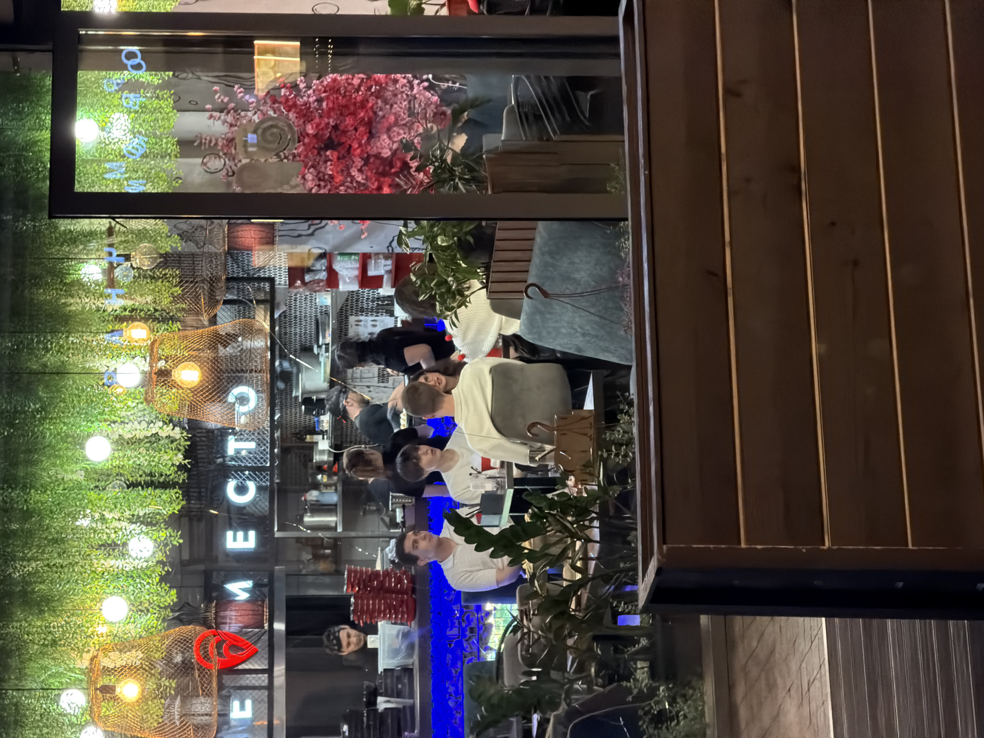 The image, taken at night, captures the reflection of a restaurant in a window. The restaurant appears busy, with multiple customers seated and dining. Some staff members can be seen in the background, presumably preparing food in the kitchen. The interior of the restaurant is decorated with plants and lights, including overhead cage-like lamps, adding to the ambiance. The exterior of the building features a brown wooden wall, and there is vegetation visible outside. The reflected text appears to read "Mesto", which translates to "place" in Russian, implying that the restaurant is potentially located in Moscow, Russia.