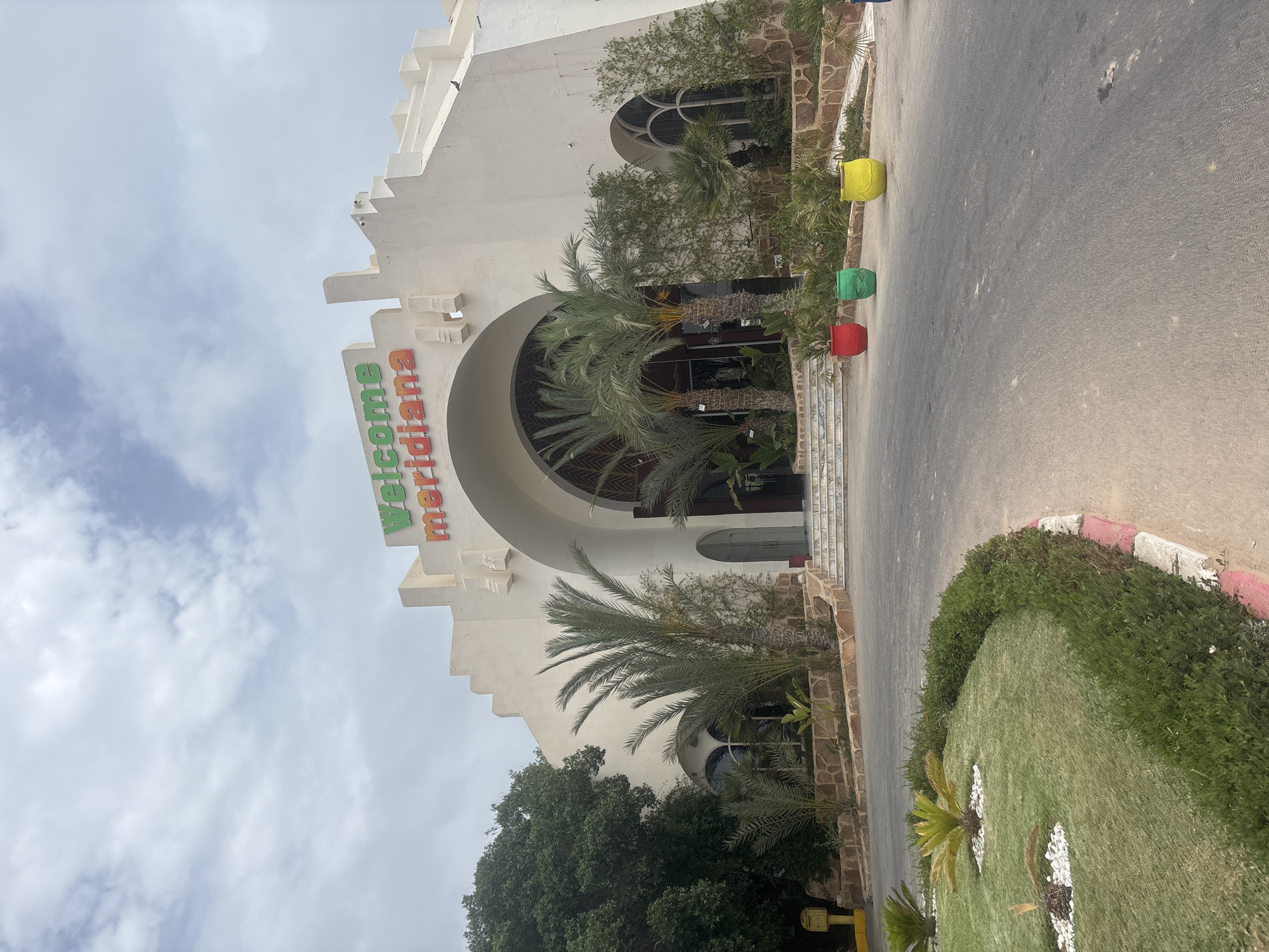The image shows the entrance to the Meridian hotel in Taguermess, Tunisia. The hotel is a large, white building with an arched entryway. Above the entrance, the words "Welcome Meridiana" are visible in green and red lettering. Palm trees are planted in front of the hotel, and there are several potted plants in colorful containers placed along the driveway. The sky is cloudy.