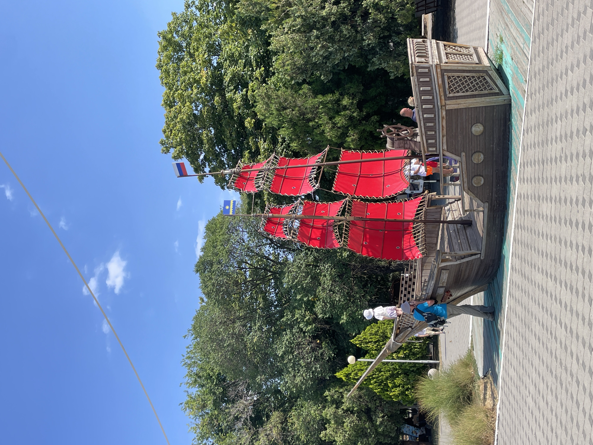 This is a daytime, eye-level, outdoor photograph of a wooden pirate ship-themed playground structure in Anapa, Russia. The structure features red sails, a wooden hull, and a steering wheel. Several people are visible on and around the ship, including a person in a blue shirt and backpack, a child in a white hat, and other individuals in the background. The playground is situated on a paved area with a checkered pattern, surrounded by green trees and a blue sky with some clouds. The overall scene depicts a recreational area on a sunny day.