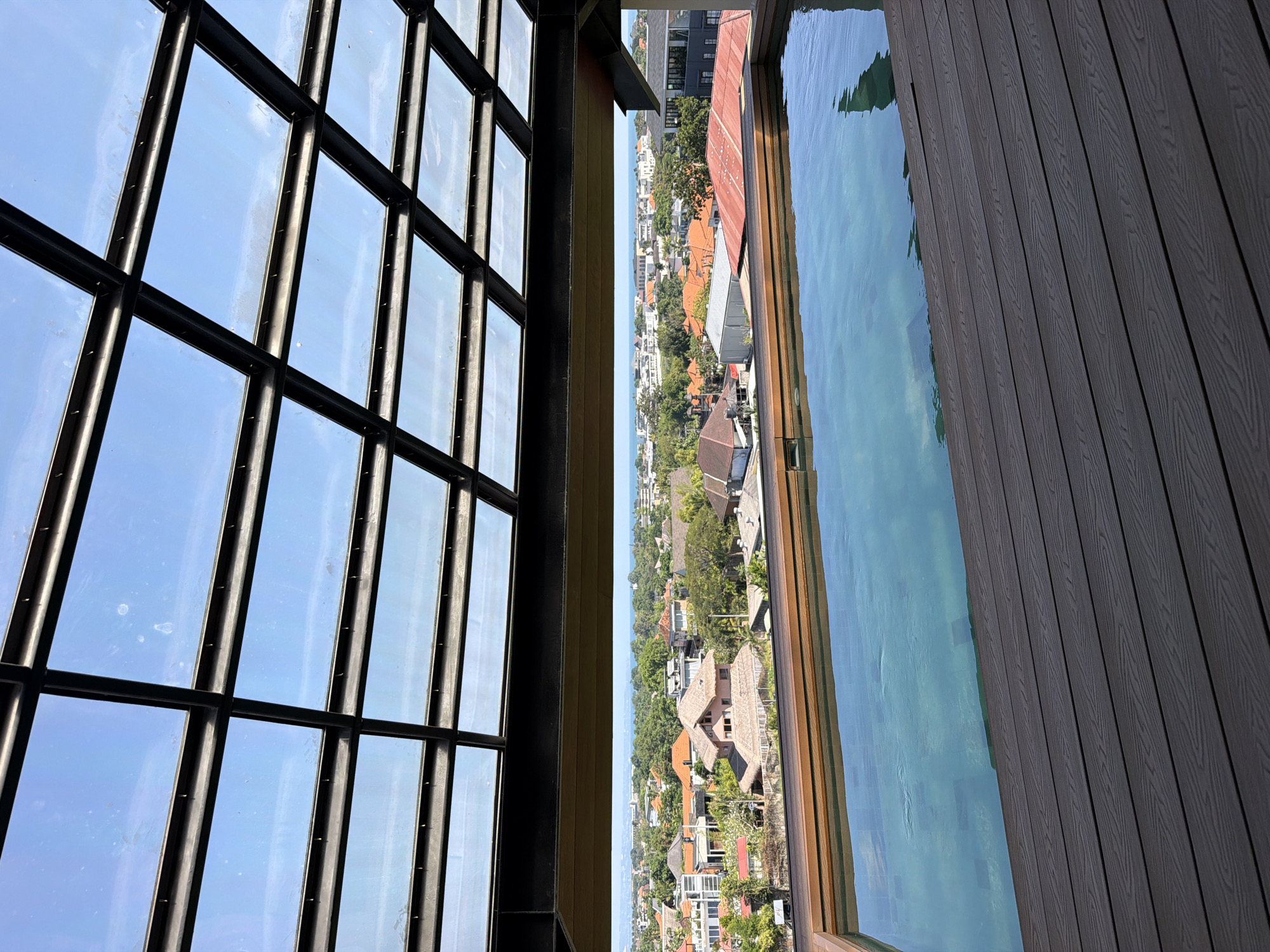 The image captures a panoramic view from a rooftop pool area in North Kuta, Indonesia, under a clear blue sky. In the immediate foreground, a dark gray, textured composite deck with horizontal planks extends, meeting a light brown wooden trim that borders a rectangular swimming pool. The pool's light turquoise water reflects the sky and parts of the overhead structure.
Above the pool and deck, a partial glass roof structure composed of a grid of dark metal beams supporting rectangular glass panels is visible, allowing sunlight to pass through. Some minor smudges are discernible on the glass panels against the clear sky.
Beyond the pool, a sprawling urban landscape of North Kuta unfolds. Numerous buildings, predominantly houses with terracotta or reddish-brown tiled roofs, are interspersed with abundant green trees and foliage. Several structures with distinct thatched or straw-like roofs are visible among the other buildings. In the far distance, a thin blue line marks the horizon, indicating the presence of the ocean. The scene is brightly lit by natural sunlight. There are no people visible in the image.