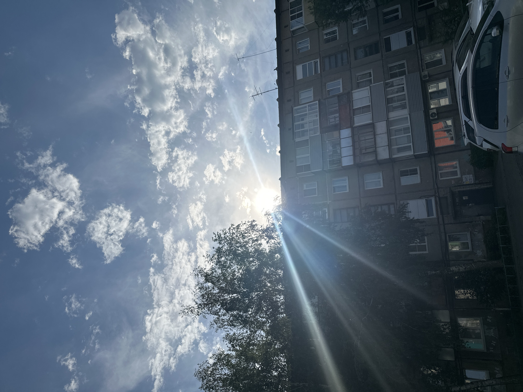 The image captures a view of a multi-story apartment building in Novoaltaysk, Russia, set against a bright, partially cloudy sky with the sun partially obscured by trees and a lens flare. The building appears to be constructed of concrete or brick, with numerous windows and balconies. Some balconies are enclosed with glass, while others are open. Various antennas are visible on the roof of the building. In the foreground, the tops of several cars are visible. Trees with dense foliage flank the building on the left side. The overall scene suggests an outdoor urban environment during daylight hours, possibly late afternoon given the angle of the sun's rays. There are no people visible in the image.