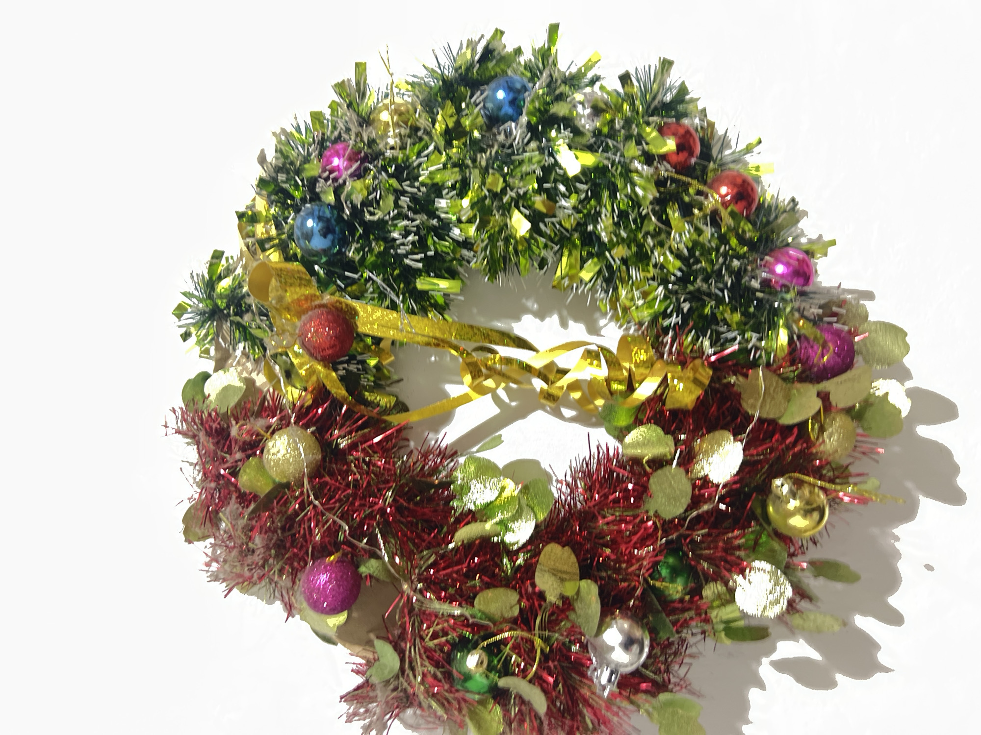 The image shows a close-up, vertical shot of a Christmas wreath hanging against a white background. The wreath is divided into two halves, one predominantly red and the other green, both adorned with tinsel, small decorative balls in various colors (red, blue, pink, gold, and silver), and metallic green leaf-like ornaments. A prominent gold ribbon weaves through the wreath. The lighting creates distinct shadows on the white surface behind the wreath, suggesting it is illuminated from the side and slightly from above. The context of Denpasar, Indonesia is not visually evident from the image itself; the scene depicts an indoor or studio setting.
