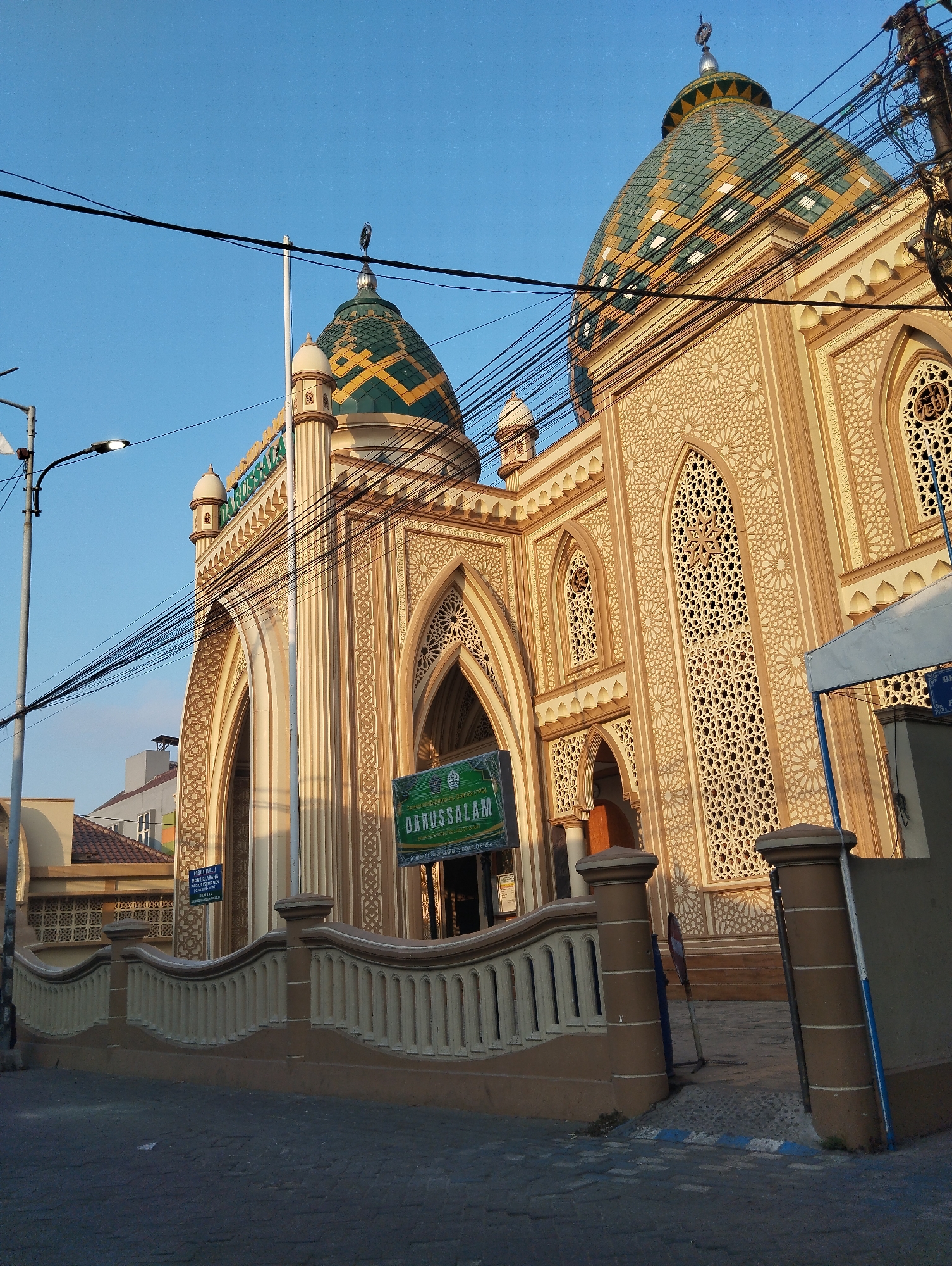 The image shows the exterior of the Darussalam Mosque in Wadungasri, Indonesia. The mosque features ornate architecture with a large, patterned green and gold dome. The building is made of light-colored stone and has intricate geometric carvings on its facade and windows. A fence with decorative pillars runs along the front of the mosque. Several utility poles and a street light are visible in the foreground, along with power lines crisscrossing the sky. The sky is clear and blue, suggesting it is daytime. No people are visible in the image.