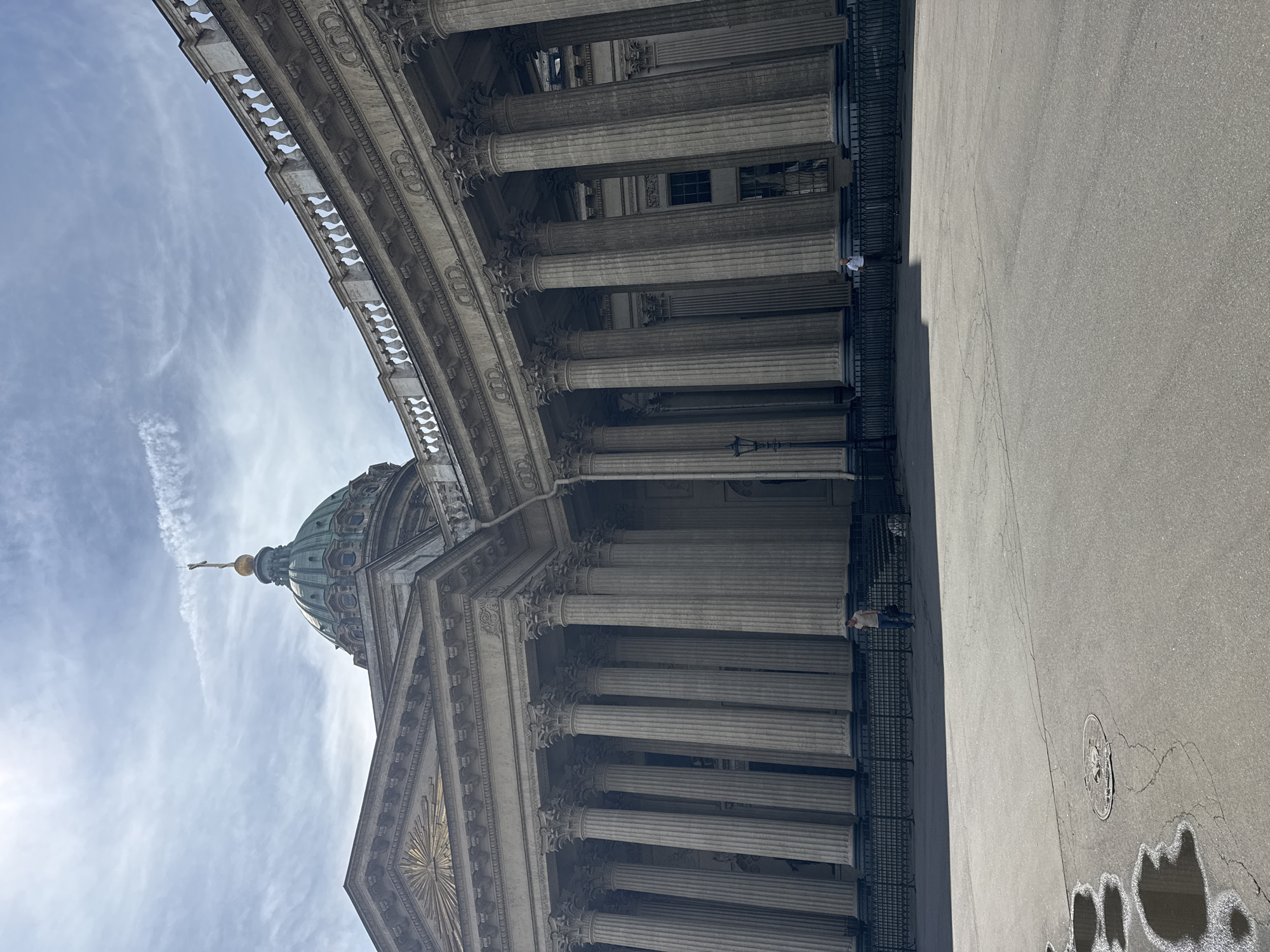 The image shows a low-angle, wide-shot exterior view of the Kazan Cathedral in Saint Petersburg, Russia. The imposing neoclassical facade of the cathedral dominates the frame, characterized by its semicircular colonnade of Corinthian columns. A green copper dome, topped with a cross, is visible against a partially cloudy sky with visible contrails.
In the foreground, a paved square is present. On the left side, there are several puddles of water, suggesting recent rain. A circular manhole cover is also visible in the pavement.
Two individuals are present in the scene. One person, wearing a light-colored shirt and dark pants, stands near the colonnade on the left, holding a dark bag. Another person, dressed in a white shirt and dark pants, stands further to the right, also near the colonnade. A black metal fence runs along the base of the cathedral. A lamppost is visible between the columns.
The lighting suggests a bright, overcast day, with the sun casting shadows from the colonnade onto the square. The overall impression is one of grand architectural scale and public space.