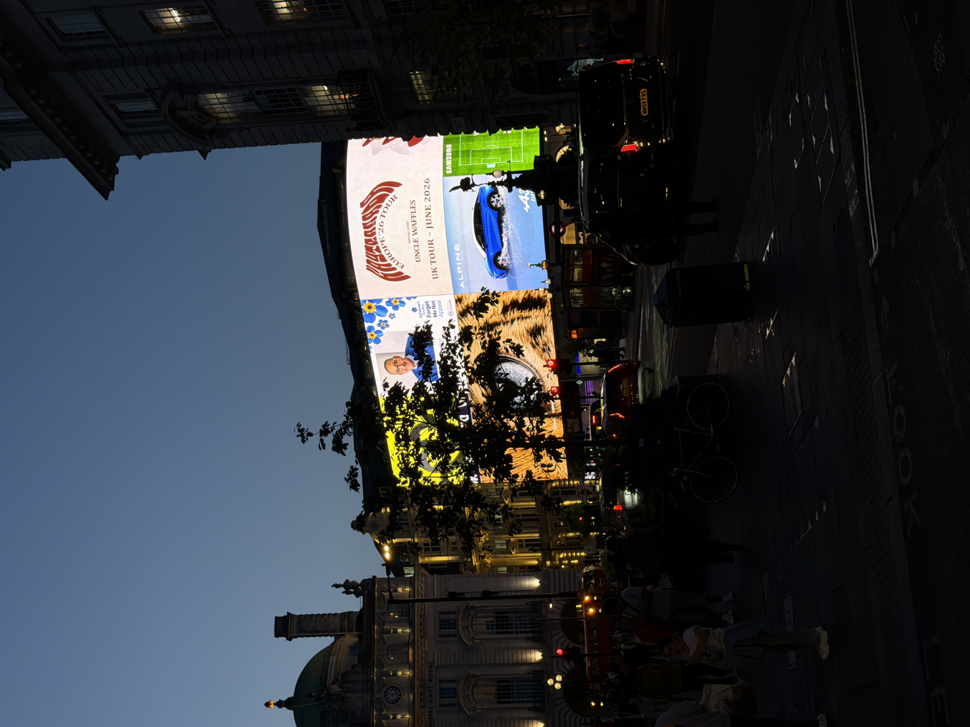 The photograph captures Piccadilly Circus in London, United Kingdom, at dusk, as indicated by the darkening sky. The iconic electronic billboards are a focal point, featuring various advertisements, including "Uncle Waffles UK Tour - June 2026" and an "Alpine" advertisement. A black cab with the registration "MM54 FRS" is parked in the street. The architectural details of the surrounding buildings are visible, including a building with a clock tower and the words "HOTEL CAFE ROYAL." A tree with sparse leaves stands in the foreground, obscuring some of the billboard display. Pedestrians can be seen on the sidewalk, their actions and interactions indistinct. The street surface is dark, and the lighting suggests it is late in the day.