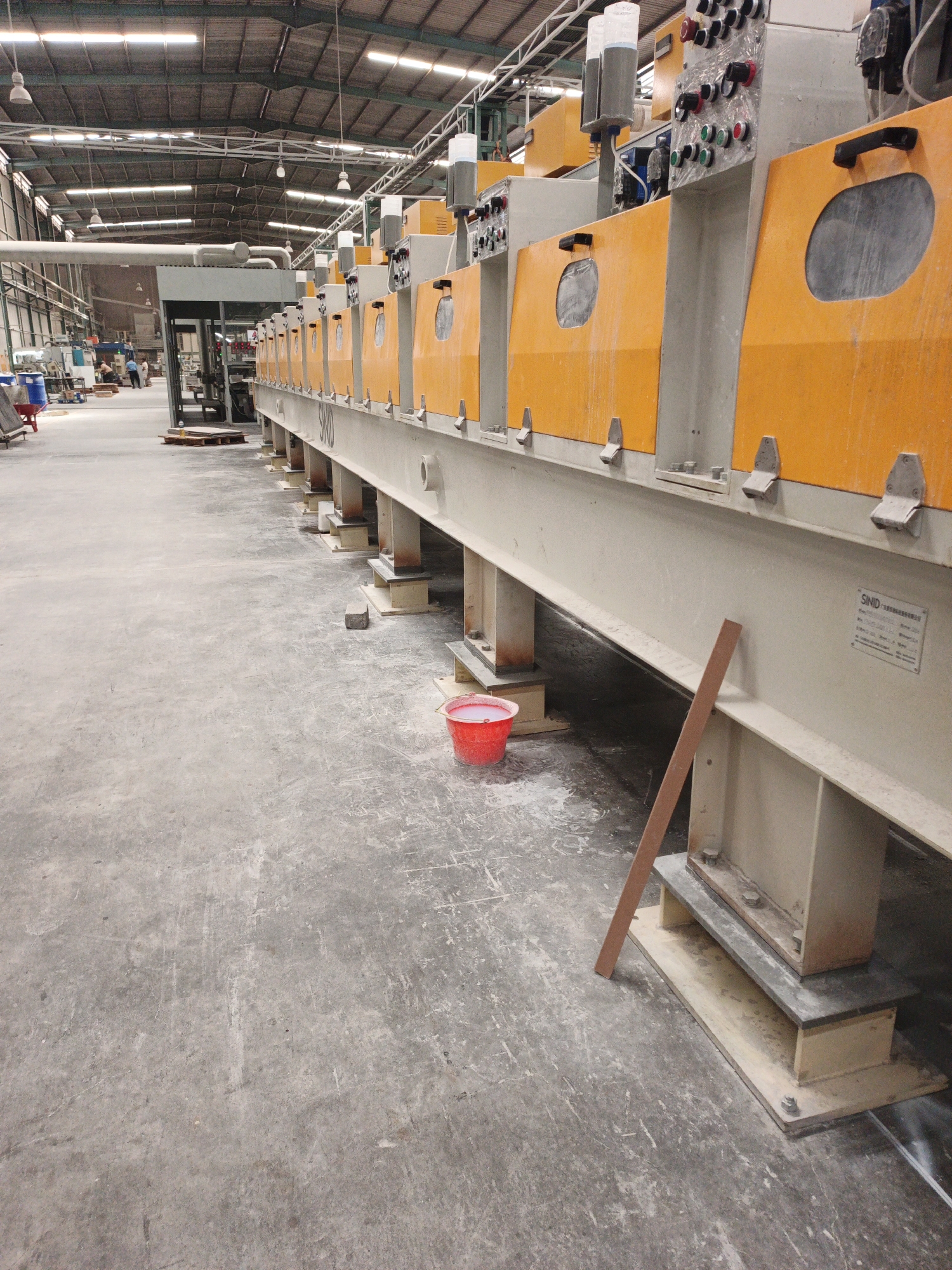The image captures a view inside a large industrial building in Ngoro, Indonesia, likely a factory or processing plant. The dominant features are a long, beige industrial machine with bright orange panels, stretching diagonally across the frame from right to left. This machine appears to be a cutting or polishing line, with multiple sections, each featuring an oval opening and a handle. Above the machine, a complex control panel with various buttons and indicators is visible.

The environment is a concrete floor that appears dusty and worn, with some scuff marks and debris. To the left of the machine, a red bucket is placed on the floor. In the background, further down the building, other machinery and industrial equipment can be seen, along with a few people who appear to be workers. The ceiling of the building features exposed beams and a lighting system with multiple fluorescent lamps. The overall impression is one of an active industrial workspace.