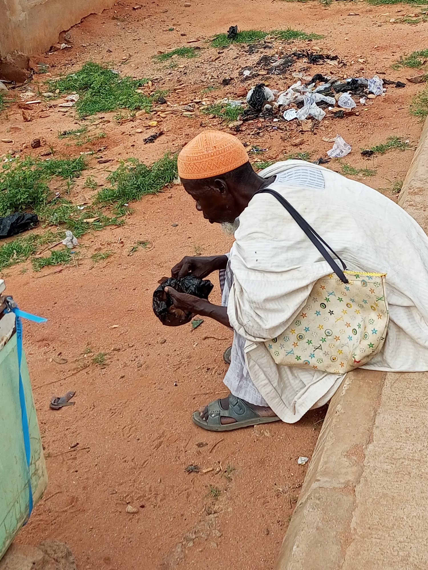 A person wearing an orange kufi, a white robe, and sandals is sitting on a concrete ledge. They are holding a black plastic bag in their hands and appear to be looking into it. The person has a beard and is carrying a patterned tote bag over their shoulder. The ground around them is sandy and reddish-brown, with patches of dry grass and scattered litter, including plastic bags and other debris. To the left of the person, a portion of a light green structure with a blue strap is visible. The scene suggests an outdoor urban or semi-urban environment, likely within the city of Gombe, Nigeria, given the typical landscape and construction materials found in the region.