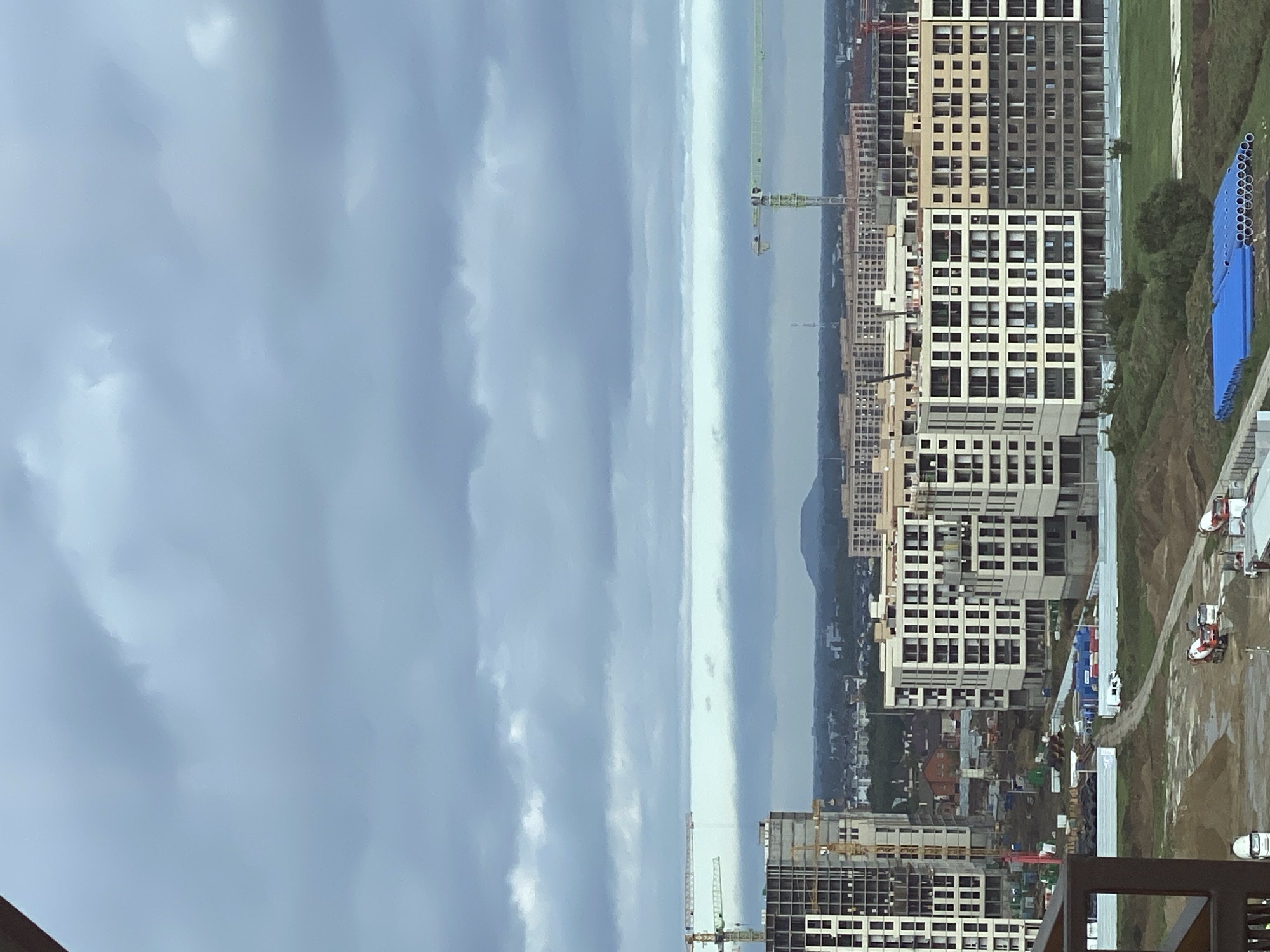 This is an aerial, medium-shot, eye-level photograph of a large construction site in the city of Krasnodar, Russia. The sky is overcast with a dramatic expanse of dark gray clouds filling the upper half of the frame, with a thin band of lighter sky visible on the horizon. In the distance, a conical mountain is visible against the hazy sky.
The foreground of the image shows the muddy terrain of the construction site, with piles of dirt and some scattered construction materials. A few concrete mixer trucks are present, one of which is actively dumping material. Blue tarps are spread out over some large pipes. A white truck is also parked on the site. The middle ground is dominated by several partially constructed apartment buildings. These buildings are multi-story, with visible concrete structures, window openings, and balconies. Yellow construction cranes are prominent, towering over the buildings and indicating ongoing development.
In the background, more residential buildings and what appear to be other construction projects stretch across the landscape. The overall impression is one of a rapidly developing urban area under a gloomy sky.