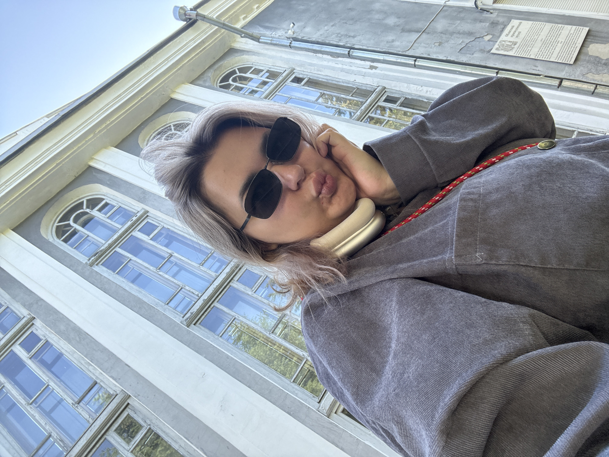 A young person wearing sunglasses and a gray shirt poses for a selfie in front of an old building with large windows in Birobidzhan, Russia. The building's facade is gray and white, with tall, arched windows. The person is leaning their head on their hand, with their lips pursed in a kiss pose. A red and green cord is visible around their neck, and they appear to be wearing a neck brace or headphones. The sky is clear and blue. The building's architectural style suggests it is likely a historical or institutional building.