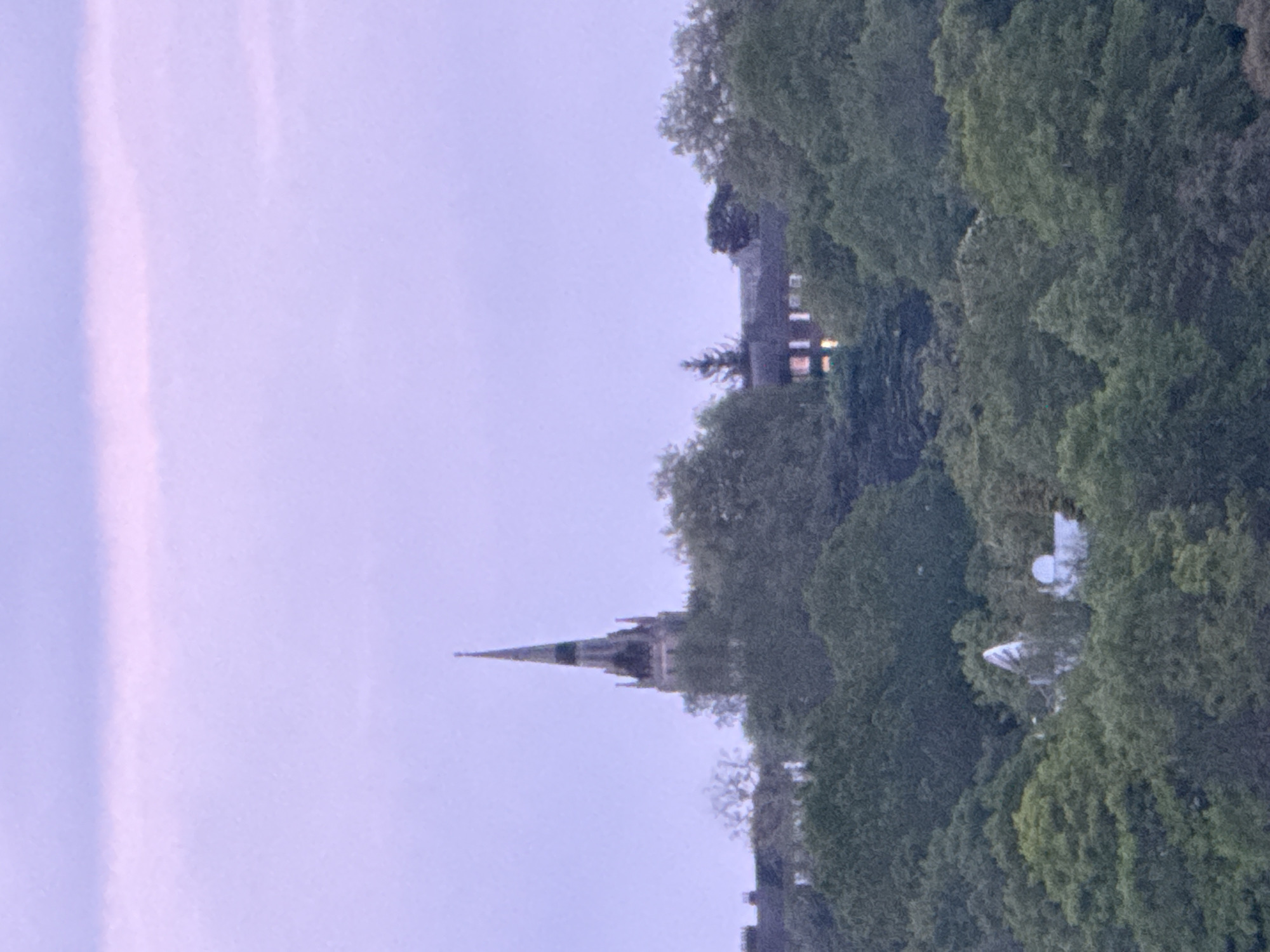 The image shows a view across trees to a church with a spire in the distance, set against a light-colored sky. The location is in the city of London, United Kingdom. There are dense green trees, with glimpses of buildings interspersed, including a residential structure. The spire of the church is prominent, and the overall scene suggests a suburban or semi-urban setting with a mixture of natural and built environments.